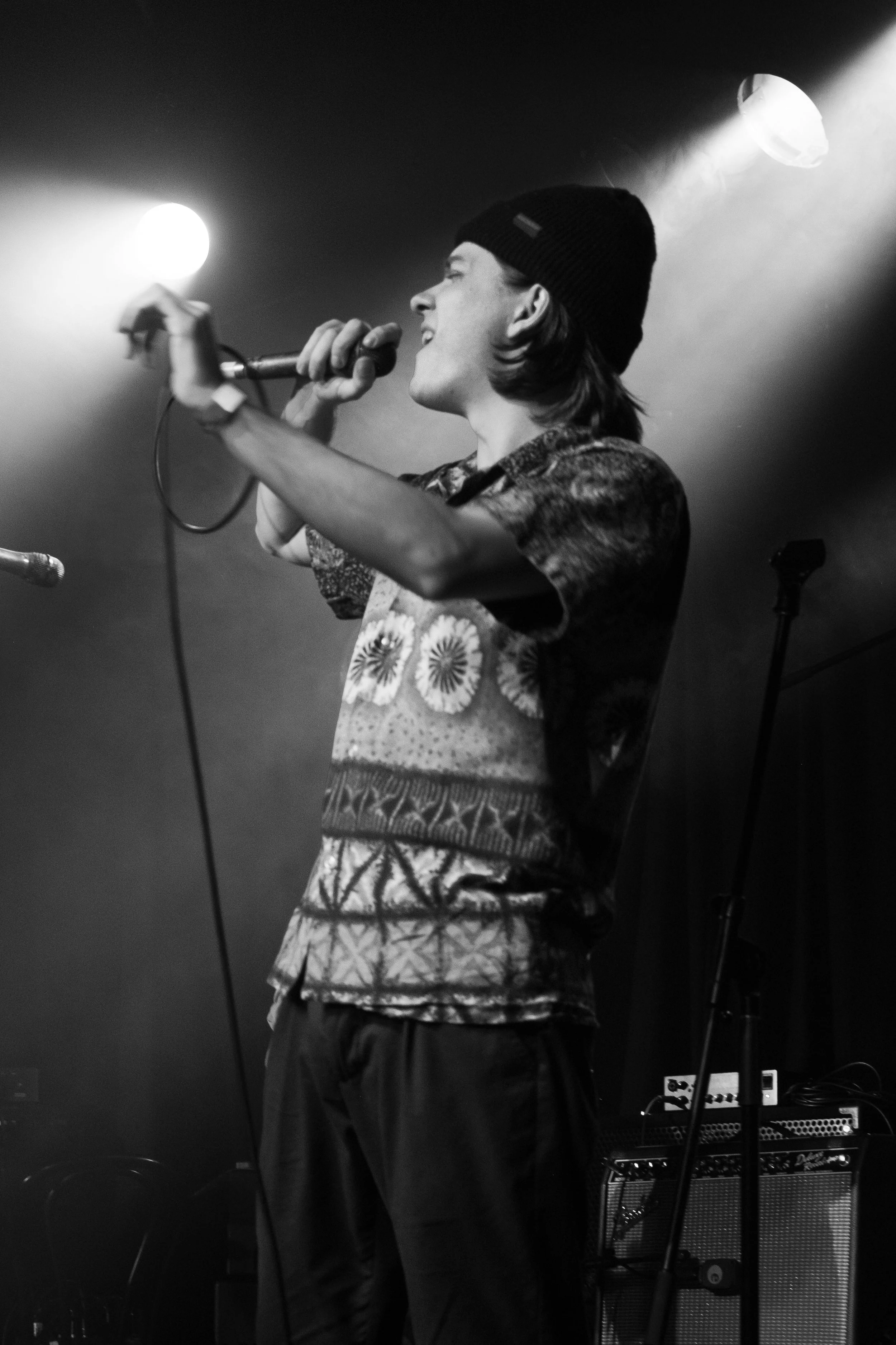 A young person performing on stage, singing into a microphone, wearing a beanie and a patterned shirt, with stage lights in the background.