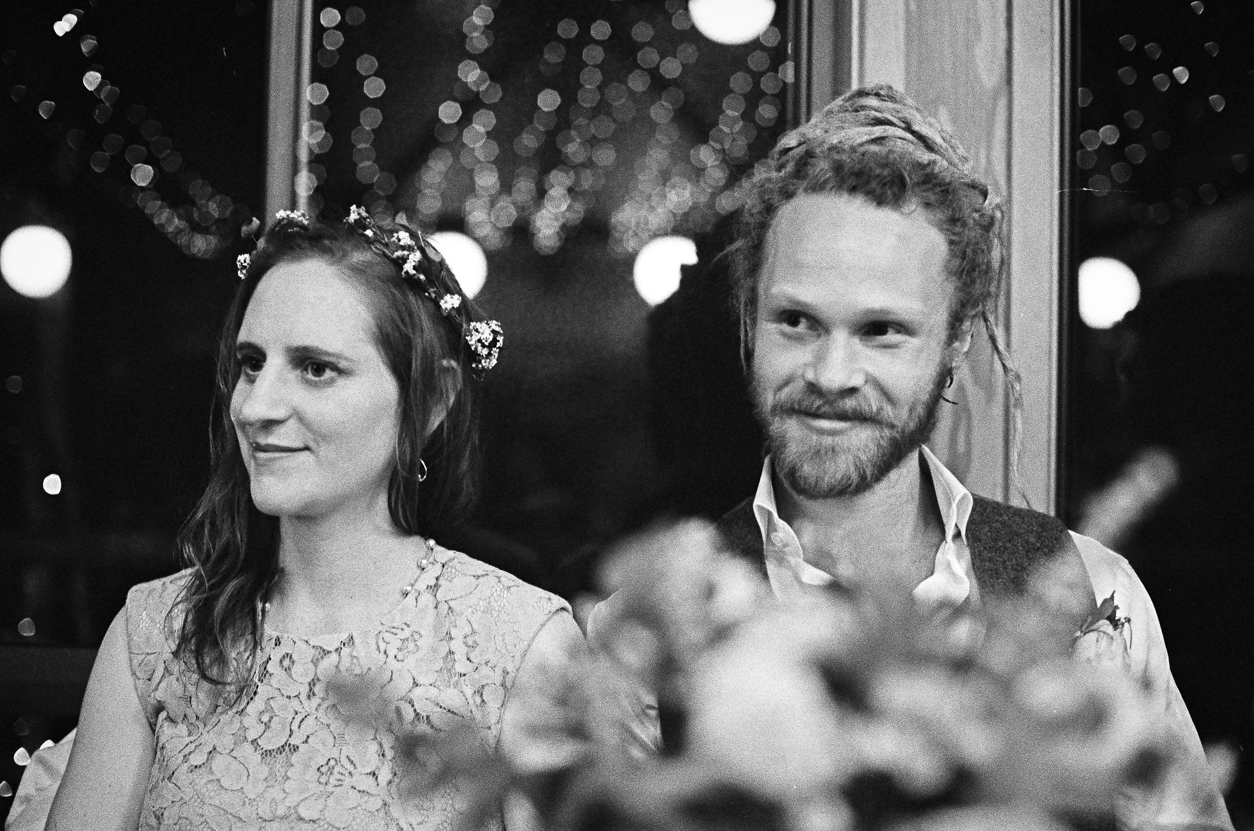 A happy couple sitting at a wedding reception, with blurred decorative lights in the background. The woman has long hair with a floral headband, wearing a lacy dress, and the man has curly hair and a beard, wearing a vest over a shirt.