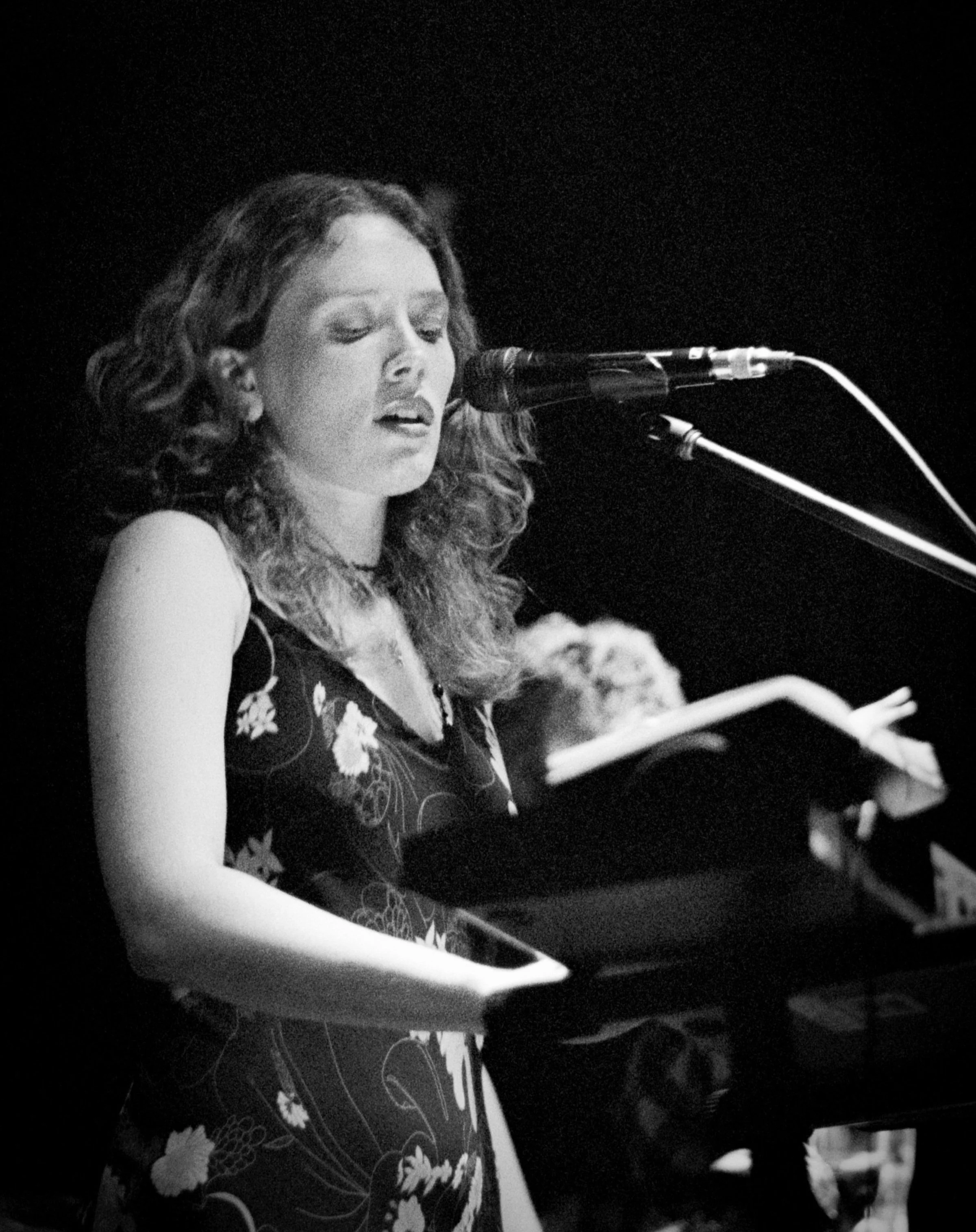 A young woman singing into a microphone while playing a keyboard on stage, wearing a sleeveless floral dress, in black and white.