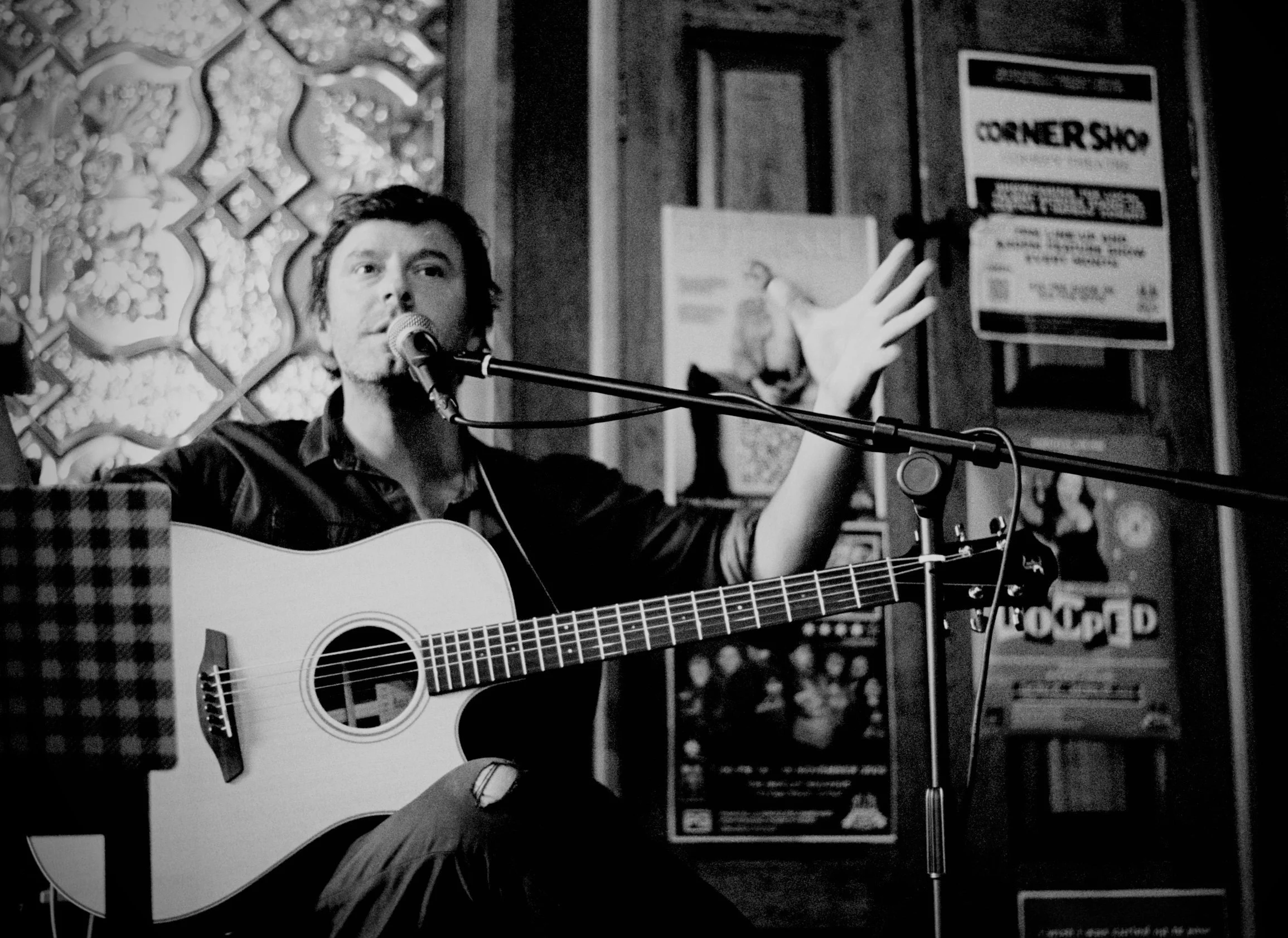 A man with dark hair performs a song while playing an acoustic guitar and singing into a microphone in a dimly lit bar or club decorated with posters and ornate designs.