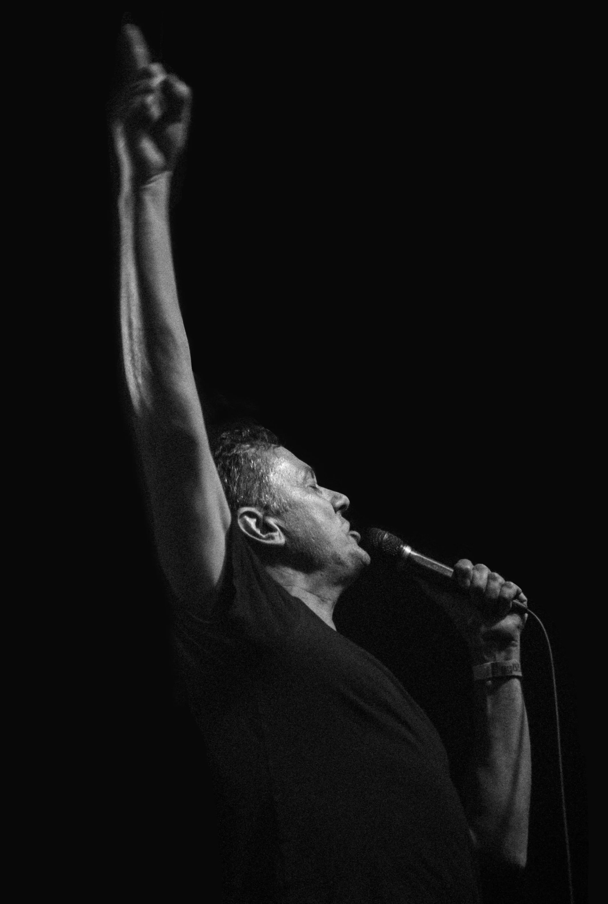 A man singing into a microphone on stage, with his eyes closed and his right arm raised, against a dark background.