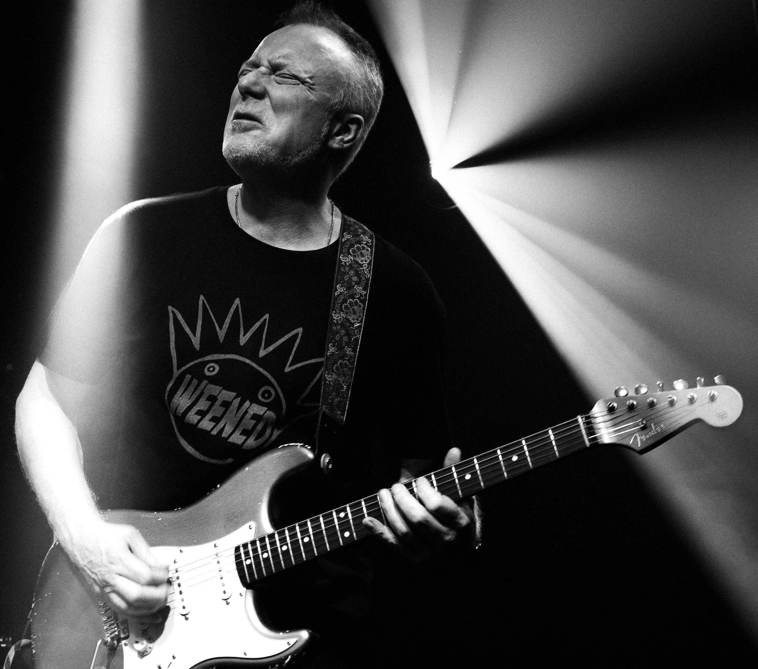 A man playing an electric guitar on stage in black and white, with stage lights in the background.