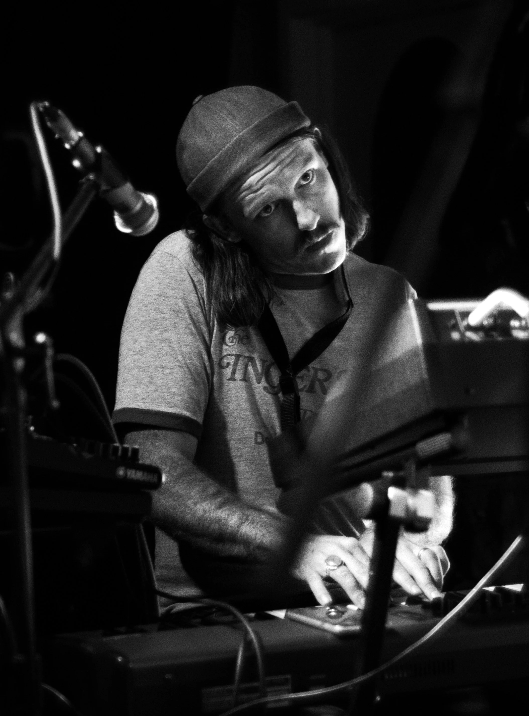 A man with long hair, a mustache, and a hat, wearing a T-shirt, playing a keyboard, with a microphone nearby, in a dark setting.
