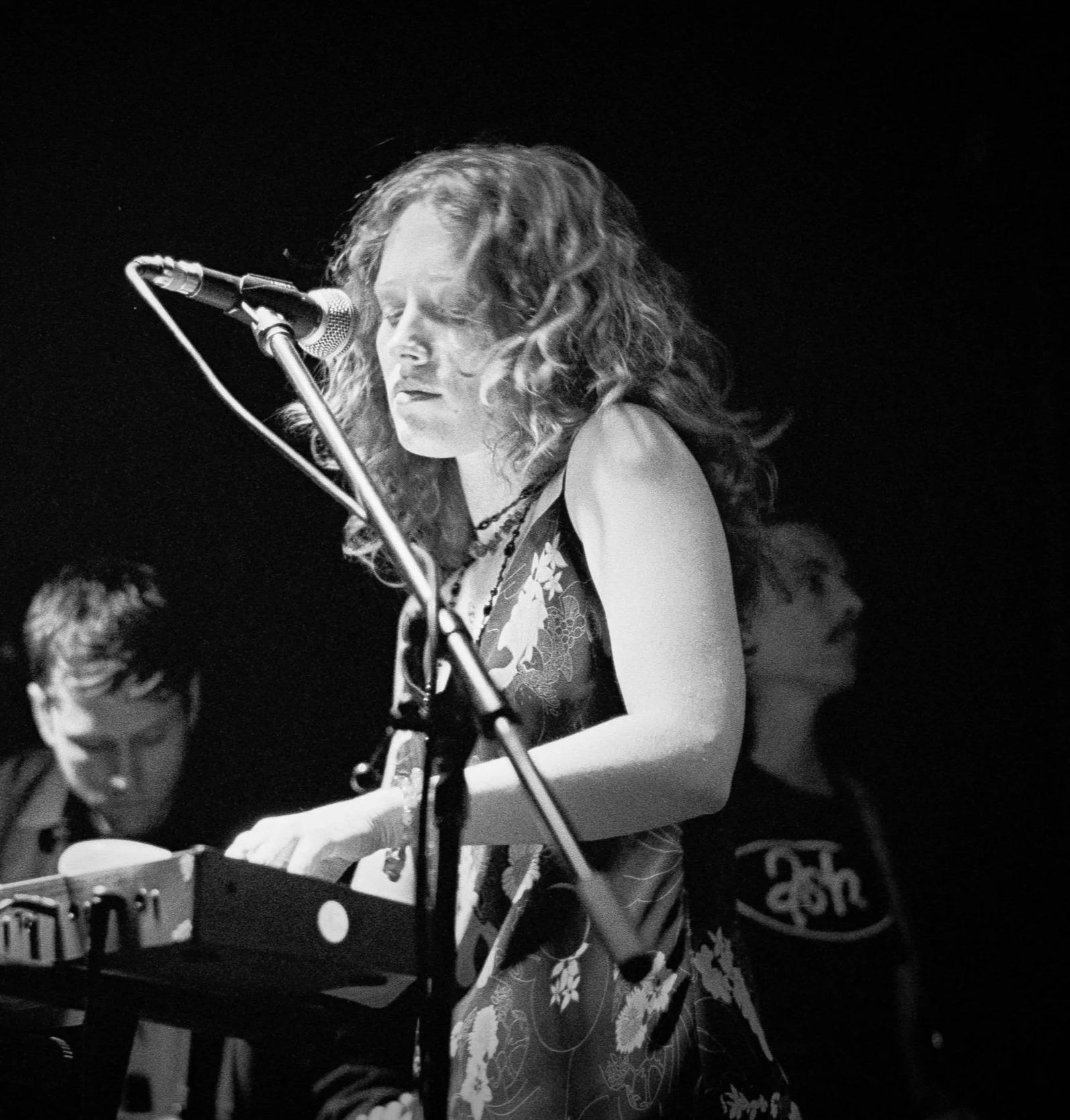 Black and white photo of a woman with curly hair singing into a microphone while playing a keyboard, with two other musicians in the background.