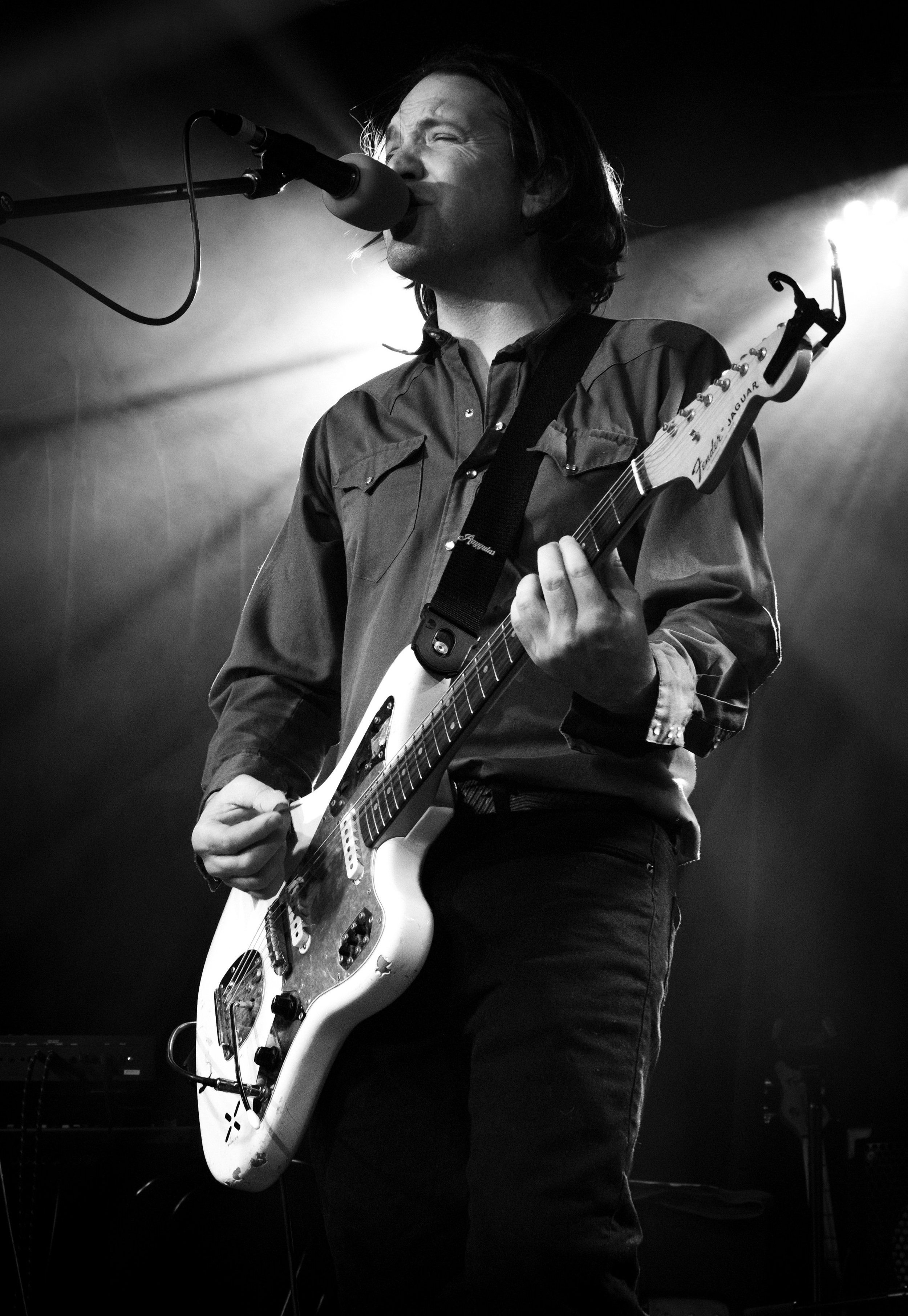 A musician with long hair singing into a microphone while playing an electric guitar on stage, black and white photo.