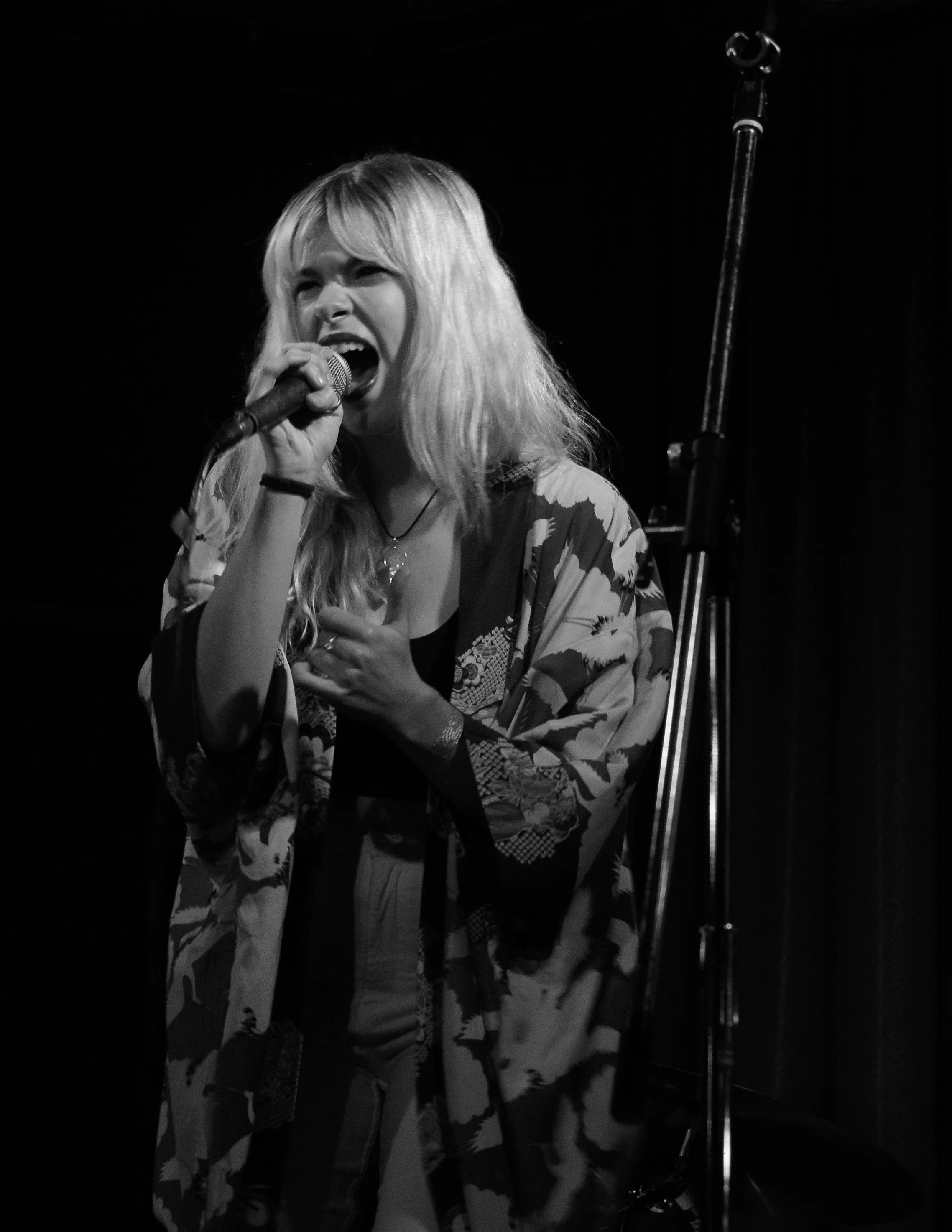 A woman with long, blonde hair singing into a microphone on stage, wearing a patterned kimono-style jacket and a black top, with a dark background.