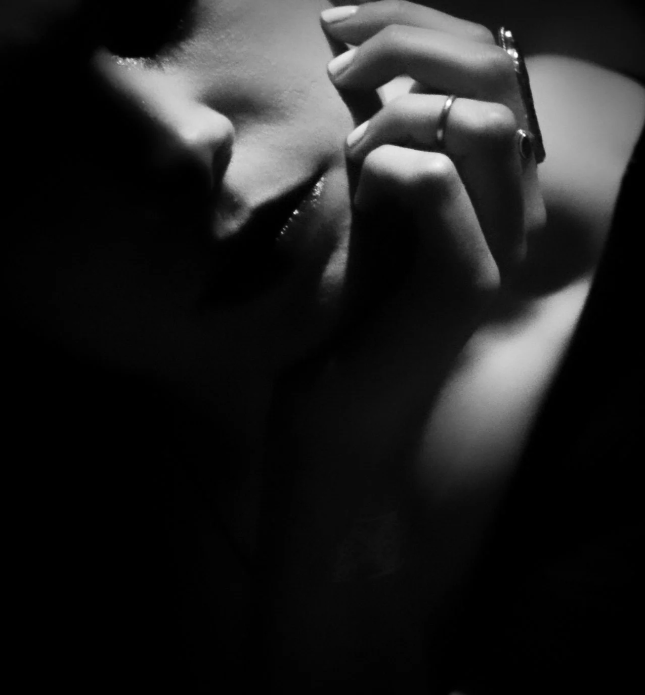A black and white close-up photograph of a woman with her eyes closed, resting her face on her hand, with her fingers near her forehead and lips, wearing rings on her fingers.