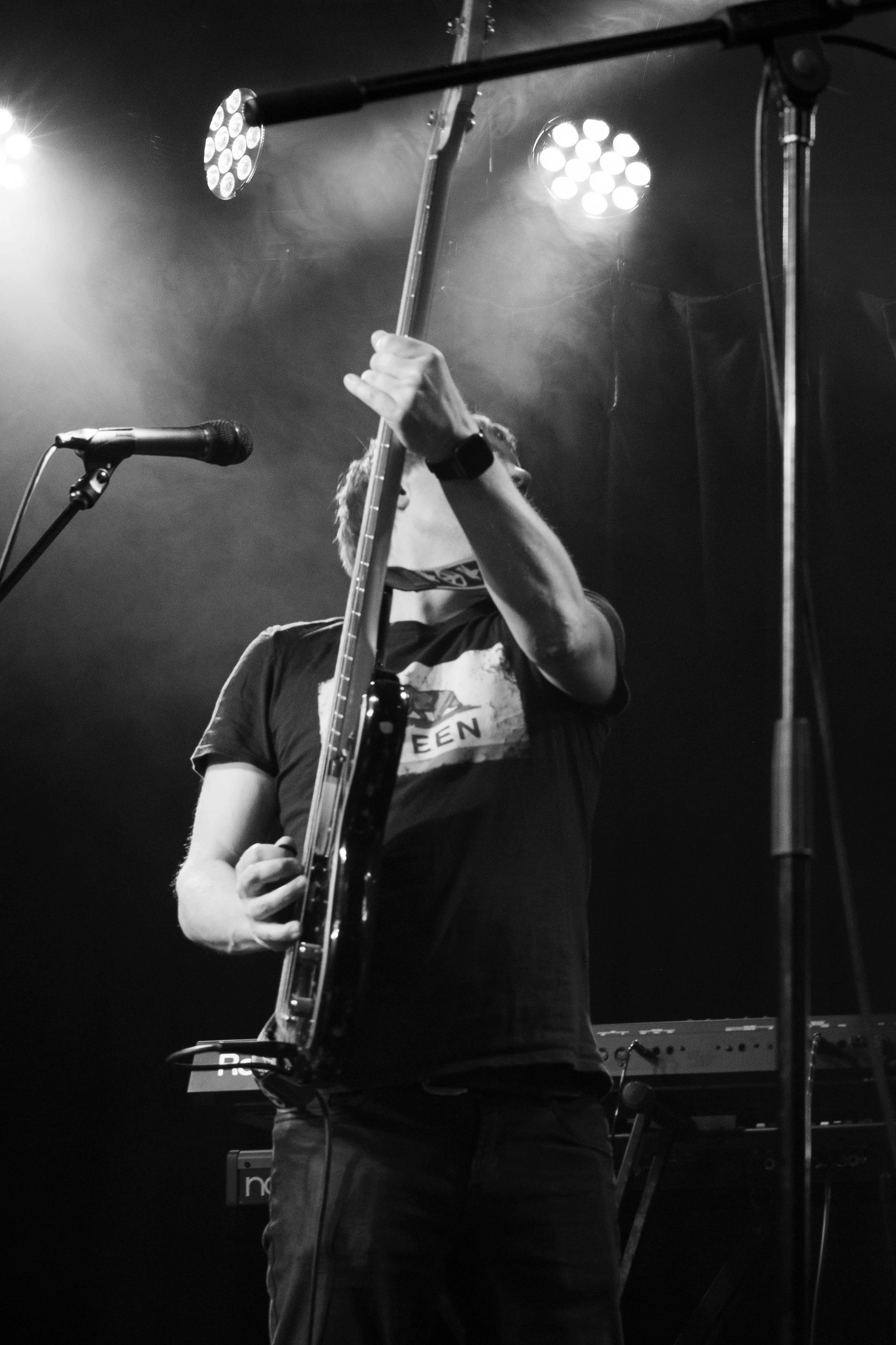 A musician on stage playing a guitar, with lighting and microphones visible, in a black and white photo.