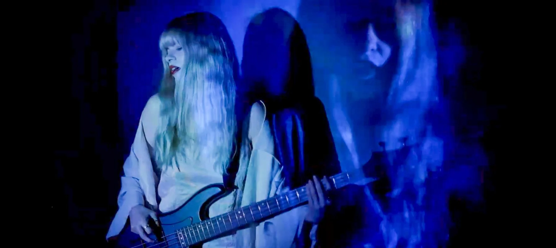 A woman with long blonde hair playing a bass guitar, singing with her eyes closed against a dark background with blue lighting.