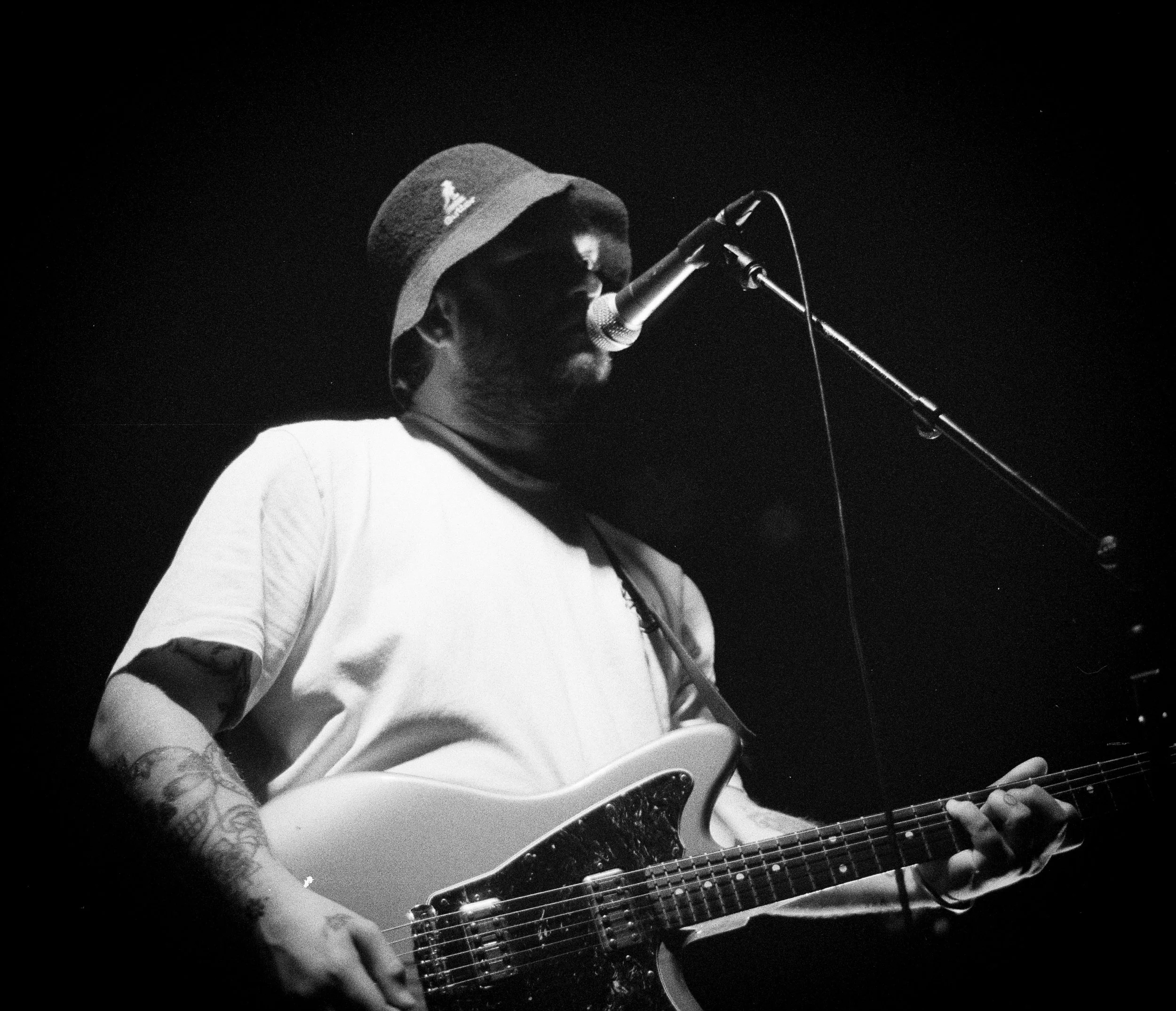 A man playing an electric guitar and singing into a microphone on stage, wearing a bucket hat and a white T-shirt.