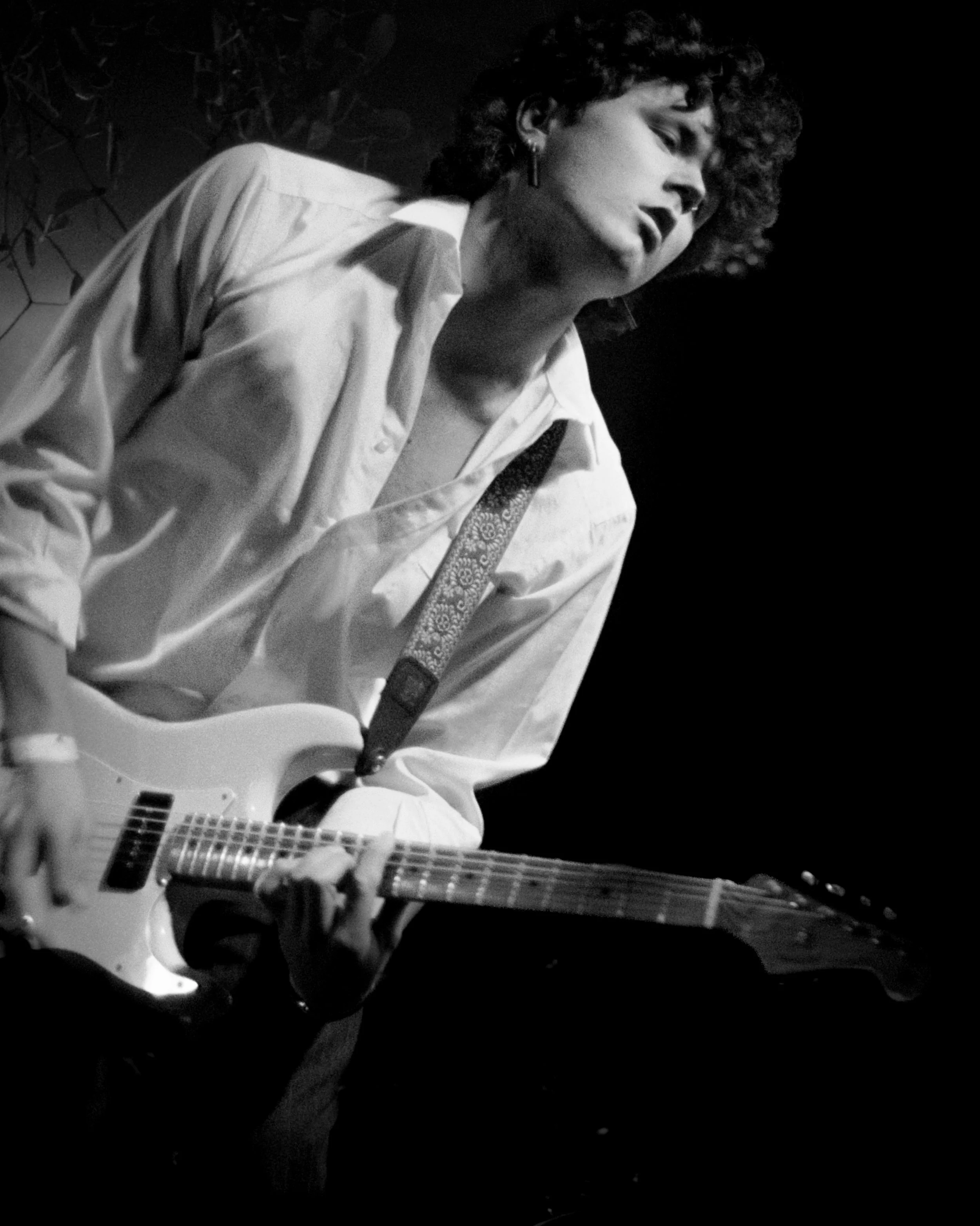 A person playing an electric guitar, wearing a loose white shirt and earrings, with curly hair, in a black and white photo.