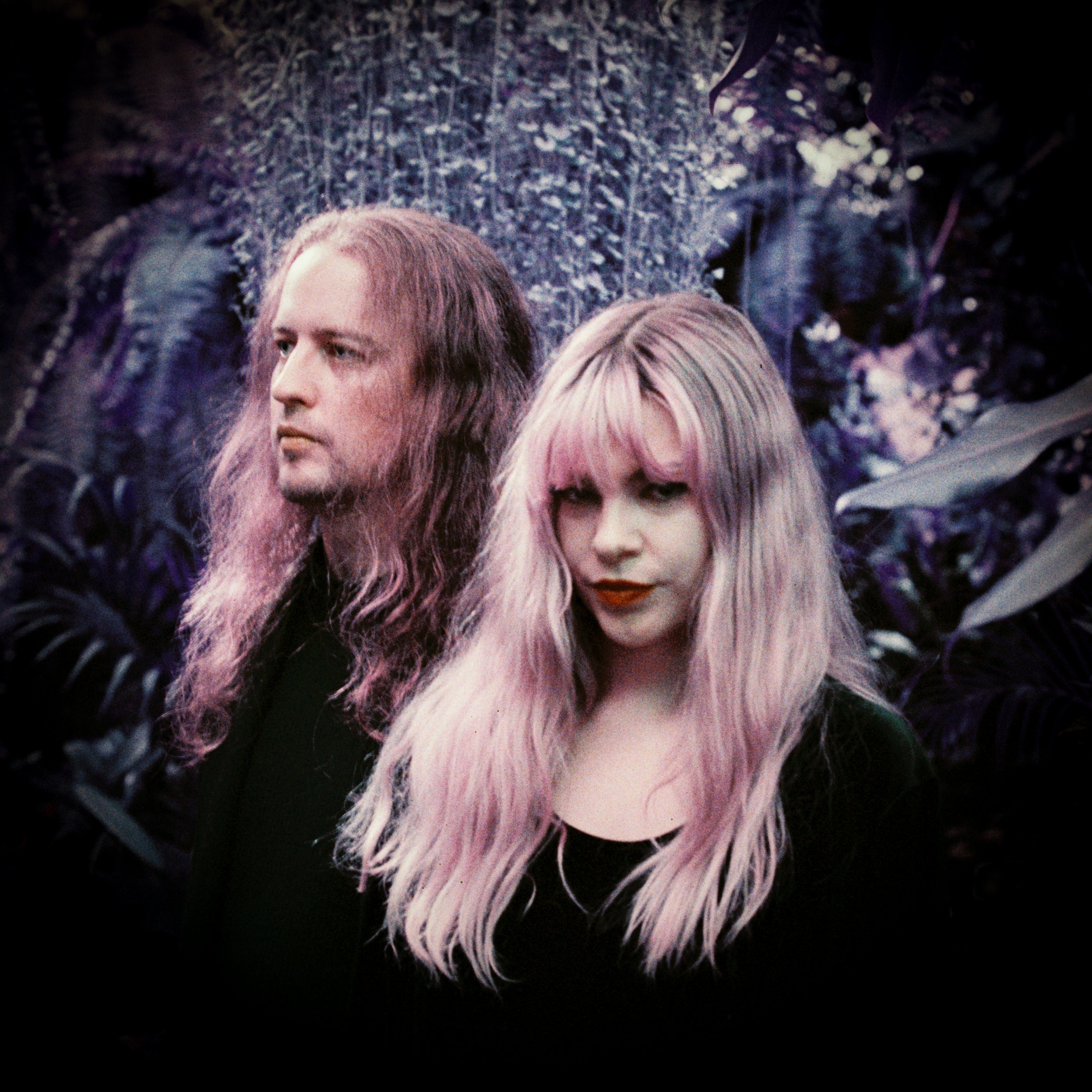 A man and a woman with pastel pink hair standing in a lush, leafy jungle background.