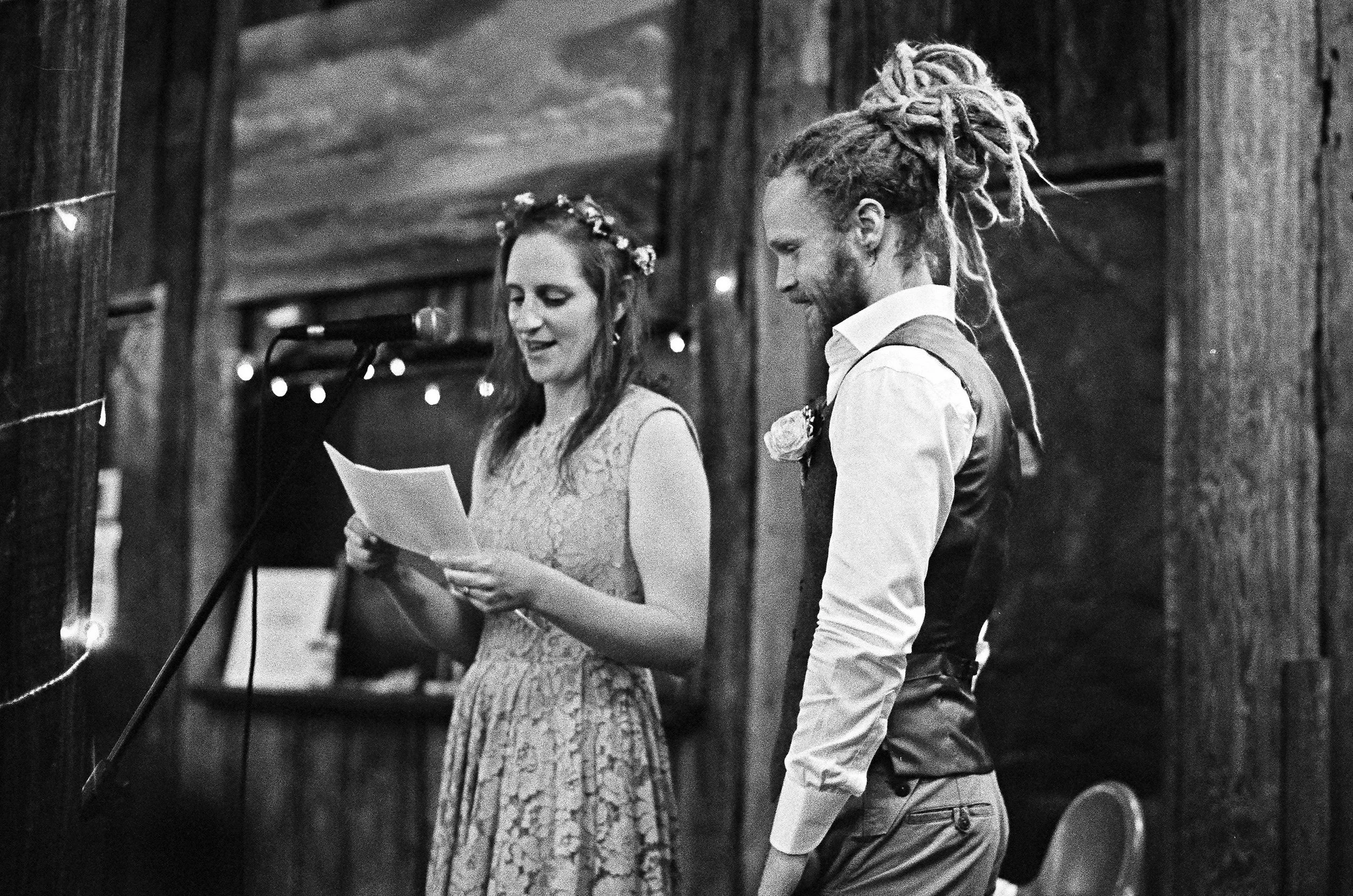A woman is reading a speech at a wedding reception, standing next to a man with dreadlocks and a vest. They are in a rustic wooden venue, and the woman is wearing a floral crown.