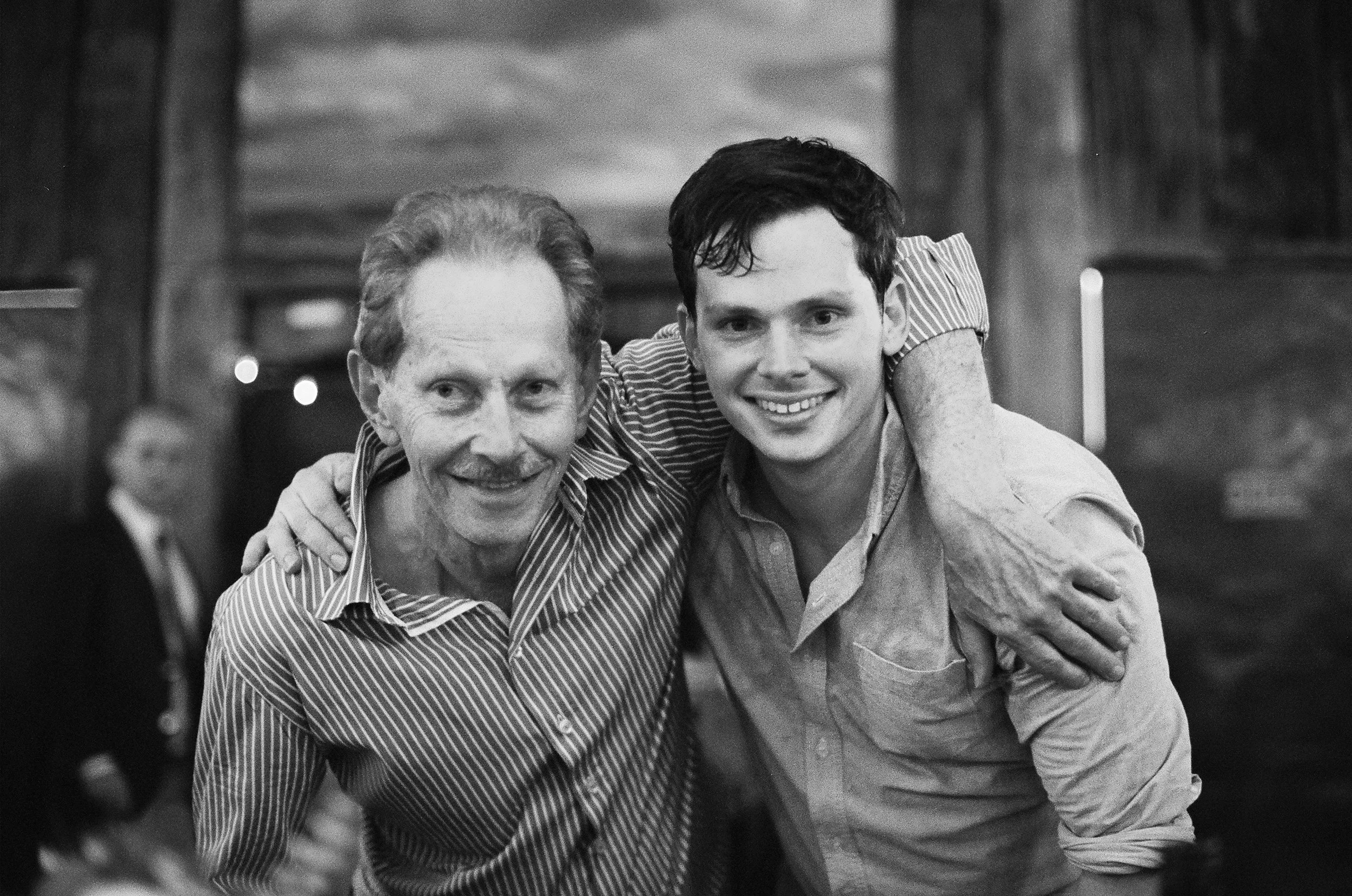 A black and white photo of an elderly man and a young man smiling and with their arms around each other in a social setting, possibly a restaurant or bar, with a blurred background.