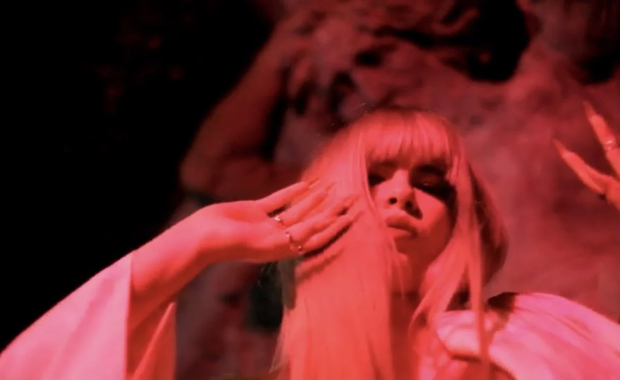 A woman with long pink hair and bangs, holding her hand to her face, appears in a red smoky environment with dramatic lighting.