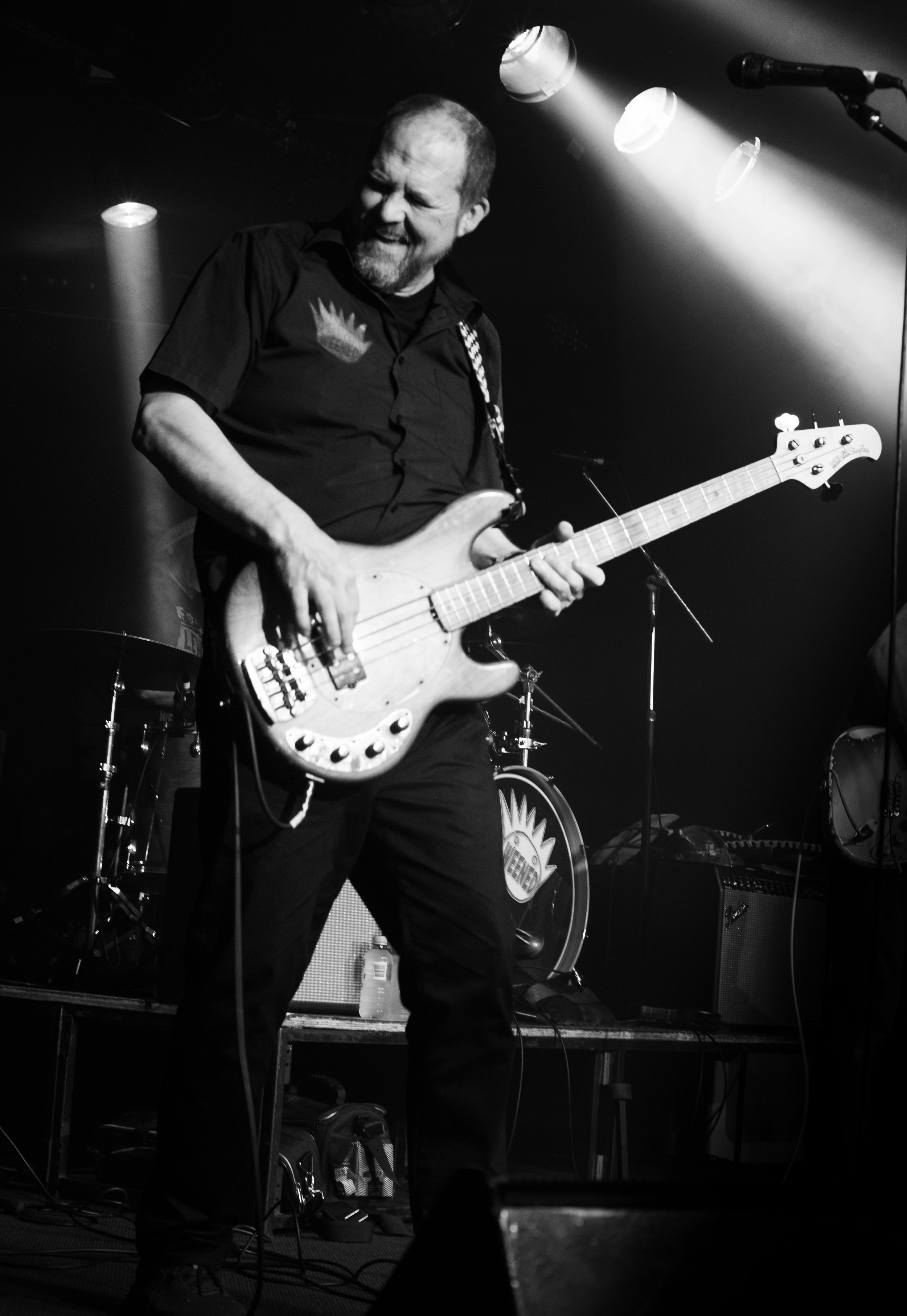 A man playing an electric bass guitar on stage during a live concert, illuminated by stage lights, with a drum set and other musical equipment in the background.