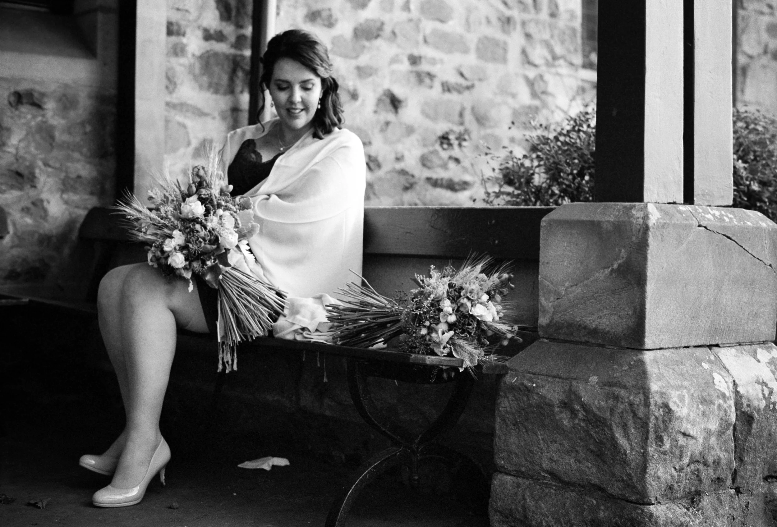 A woman sitting on a bench with bouquets of flowers around her, wearing high heels and a shawl, with a stone wall in the background.
