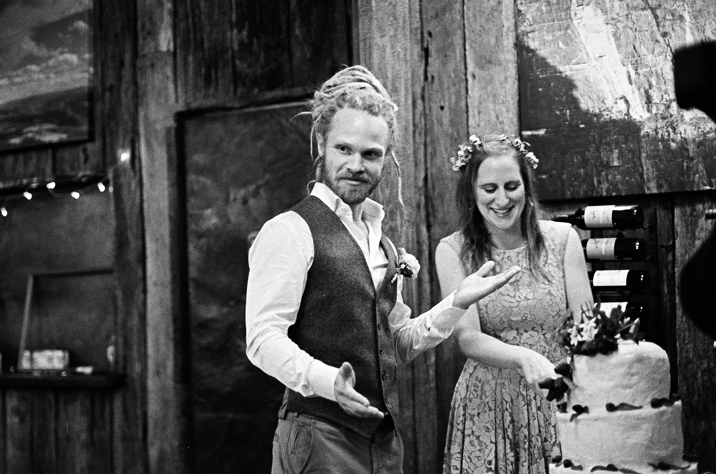 A wedding couple cutting their wedding cake, with the groom having dreadlocks and the bride wearing a floral headband, inside a rustic wooden venue.