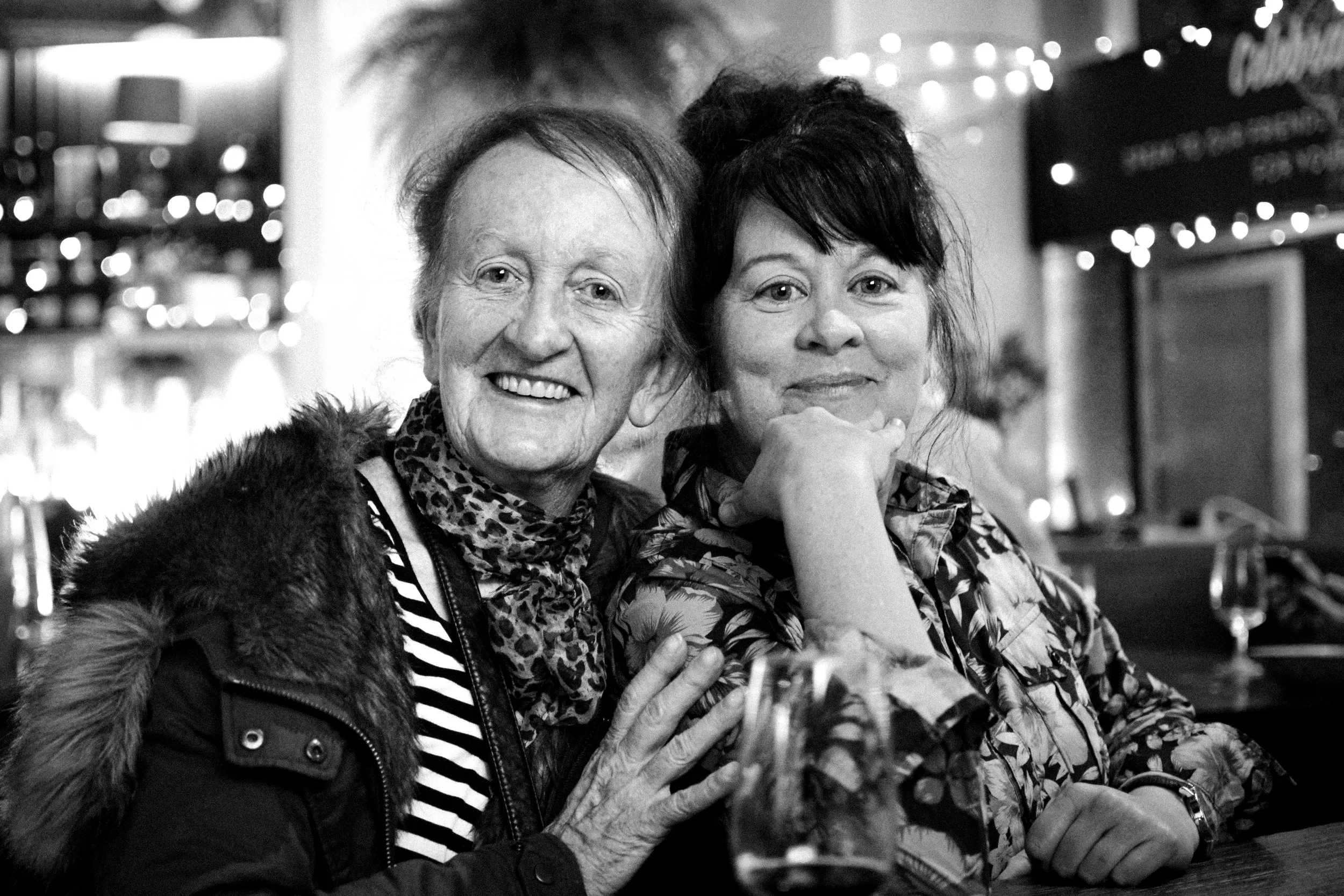 Two women smiling at a restaurant or bar, with drinks in front of them, in a cozy, decorated setting with string lights.
