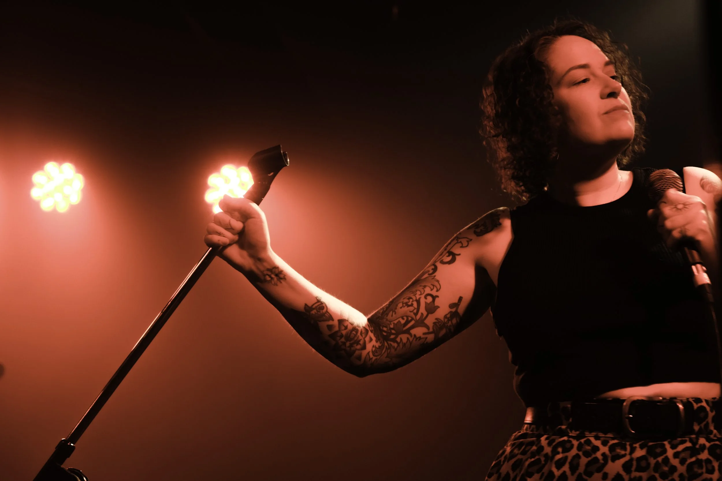 A woman with curly hair holding a microphone in her left hand and a mic stand in her right hand, with red stage lights in the background.