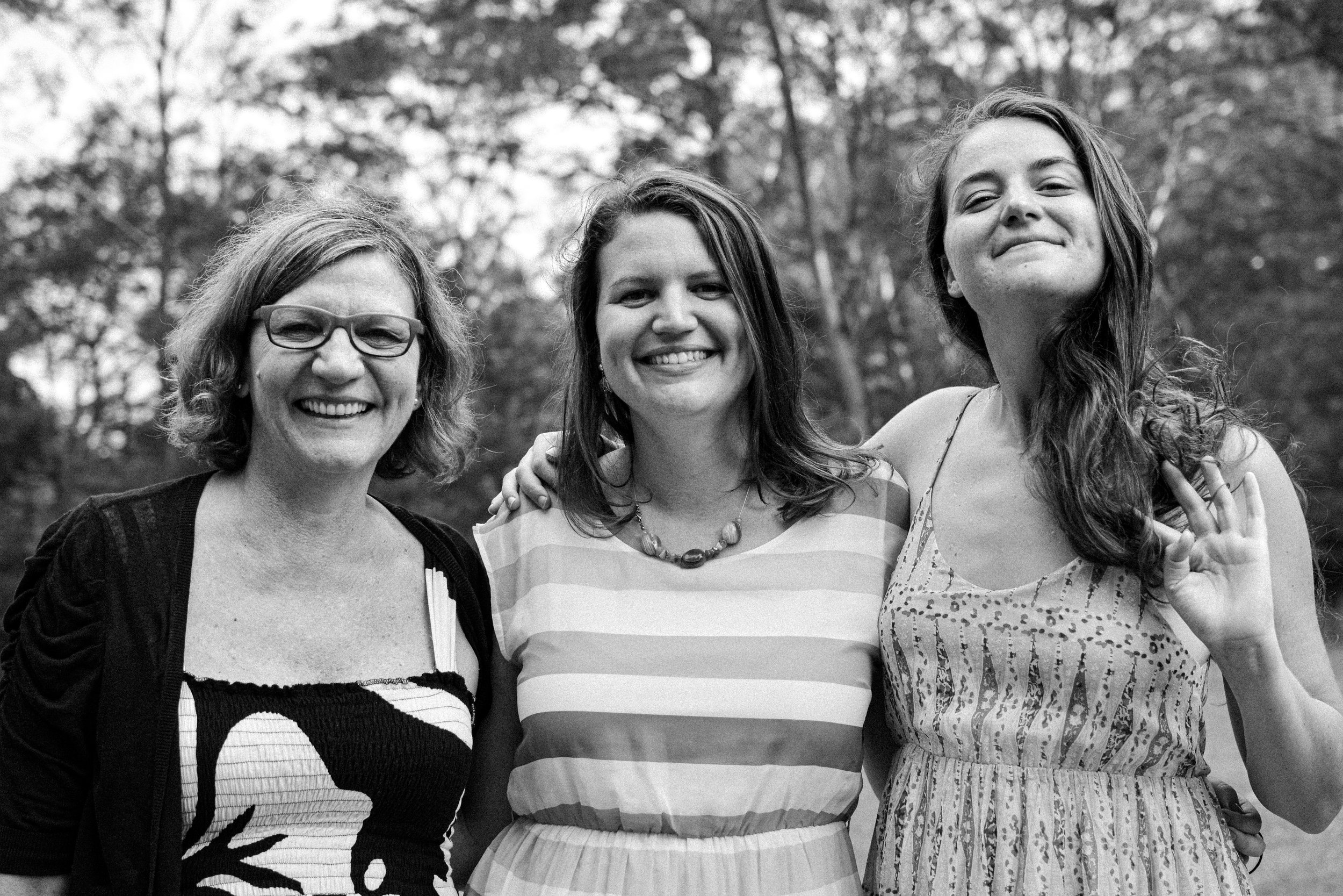 Three women smiling outdoors, one making a hand gesture, in a park or wooded area.