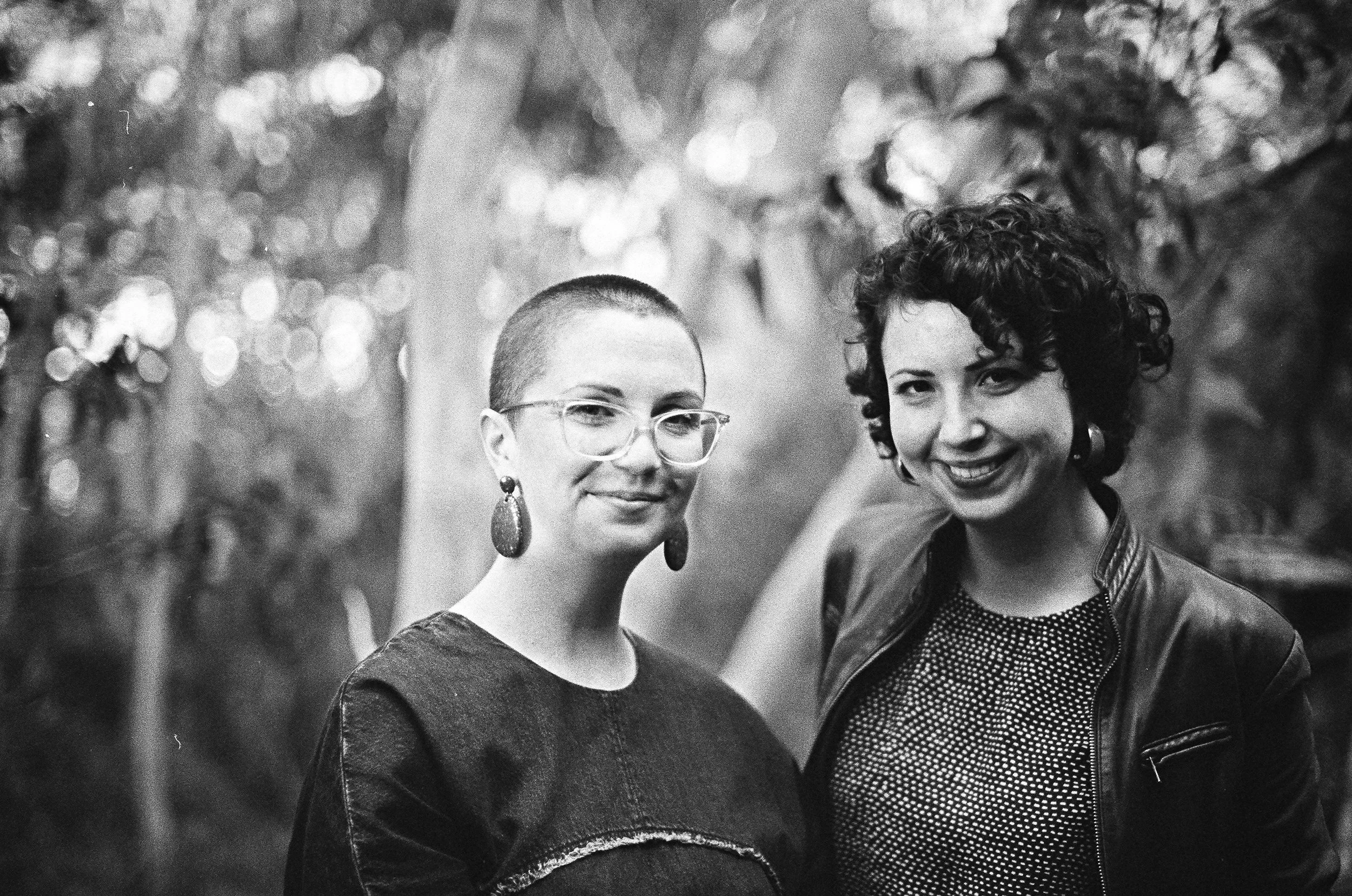 Two women standing outdoors in front of blurred trees, smiling at the camera, one with short hair and glasses, the other with curly hair and no glasses.
