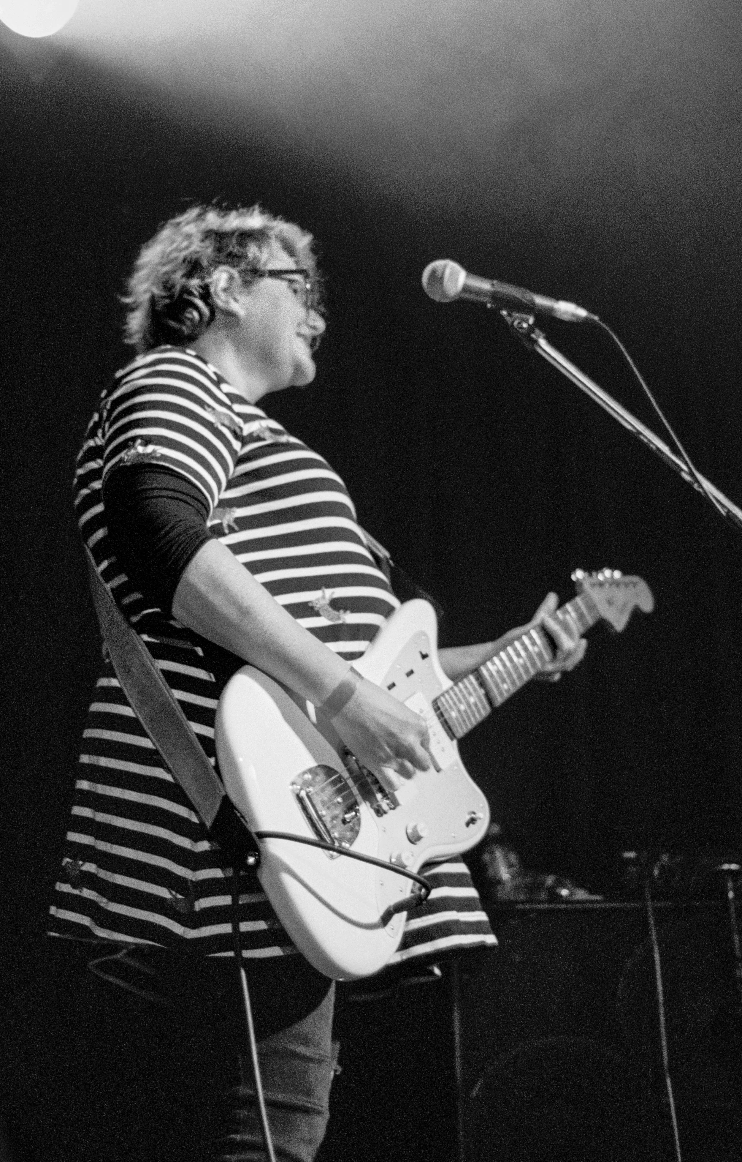 A woman playing an electric guitar on stage, wearing a striped shirt and glasses, with a microphone in front of her, black and white photo.