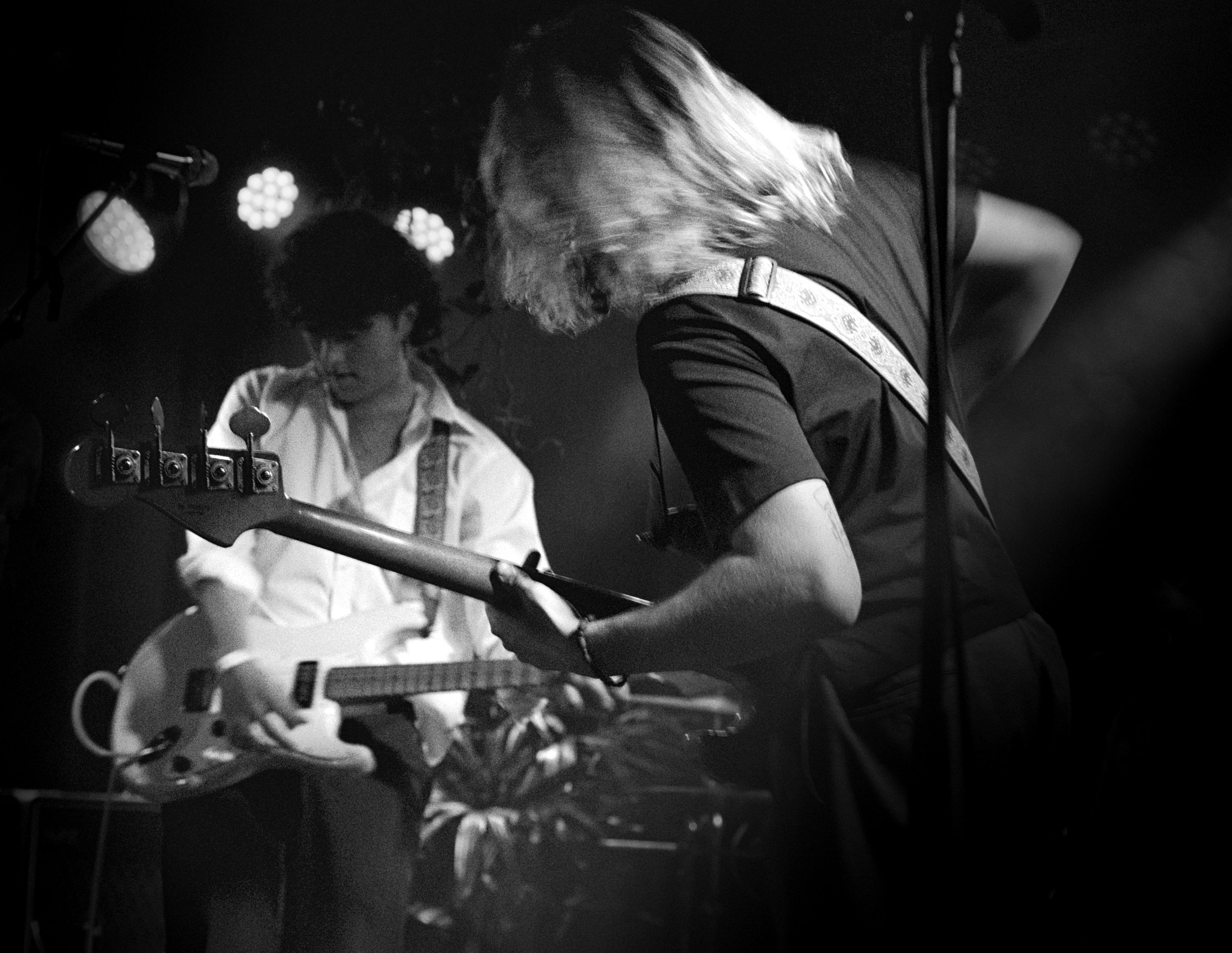 Two musicians performing on stage, one playing an electric guitar and the other playing a bass guitar, with stage lighting in the background.