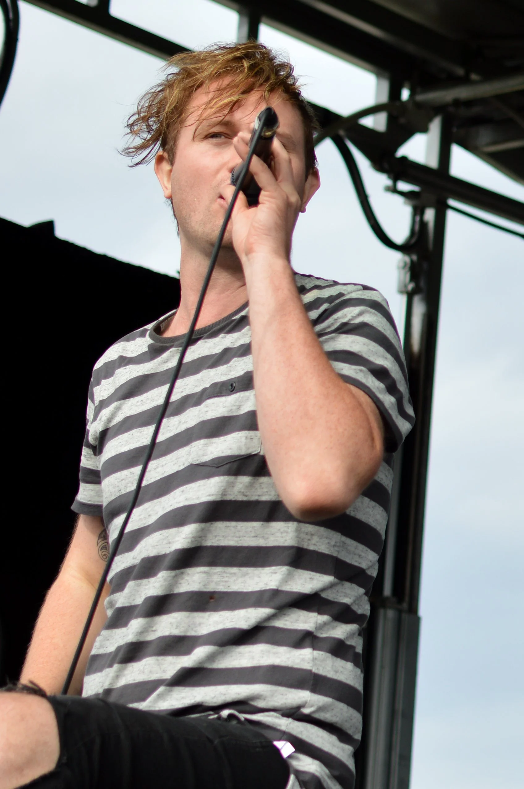 A young man with messy, light brown hair holding a microphone and singing on an outdoor stage, wearing a gray and black striped t-shirt.