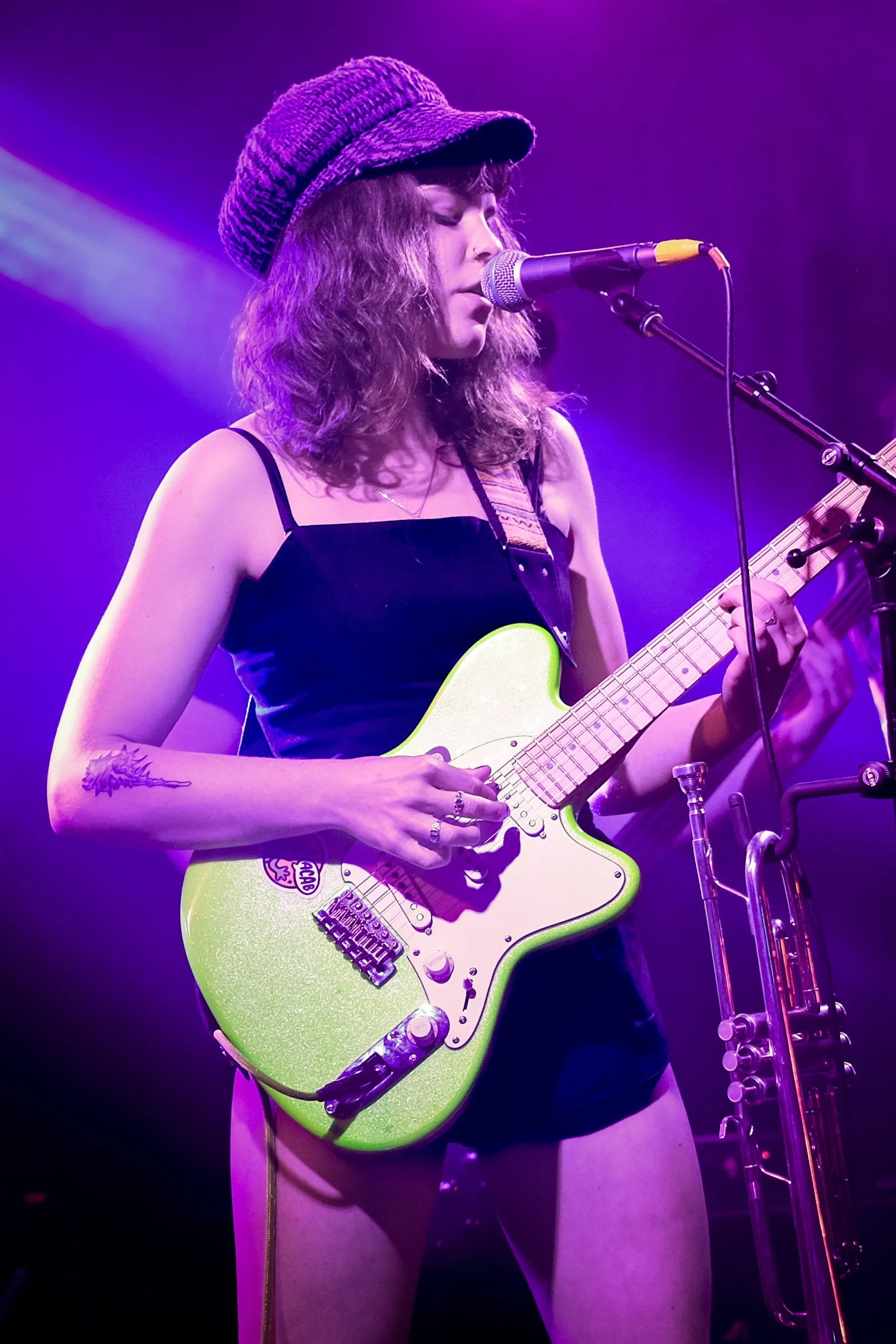 A young woman with curly hair wearing a purple knit hat, a black sleeveless top, and black shorts, playing a lime green electric guitar while singing into a microphone on stage with purple and yellow lighting.