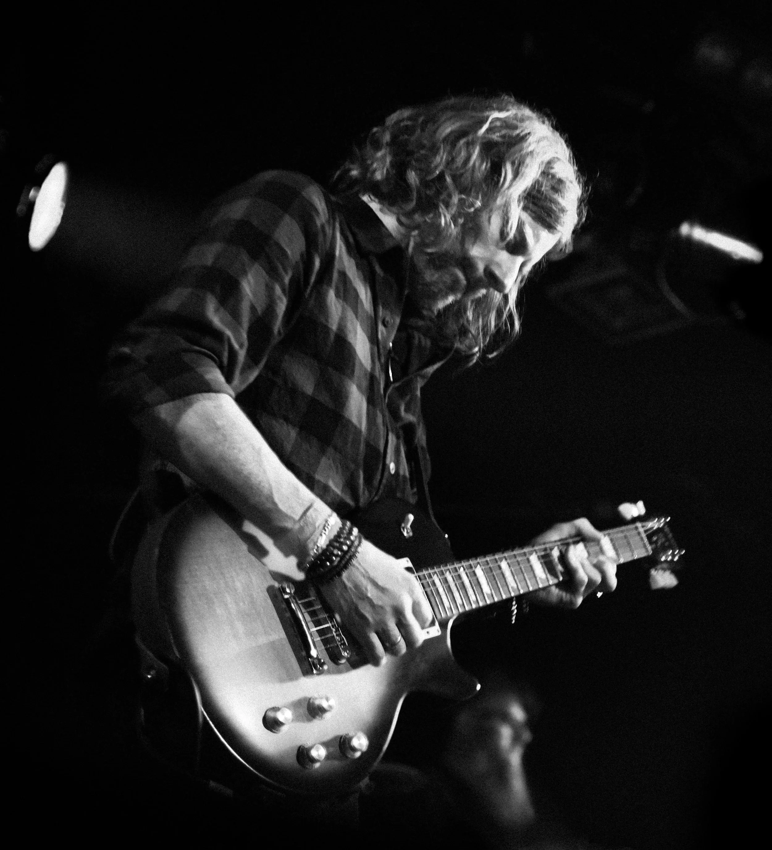 A male guitarist playing an electric guitar on stage, wearing a checkered shirt and multiple bracelets, with long hair and facial hair, in a black and white photo.