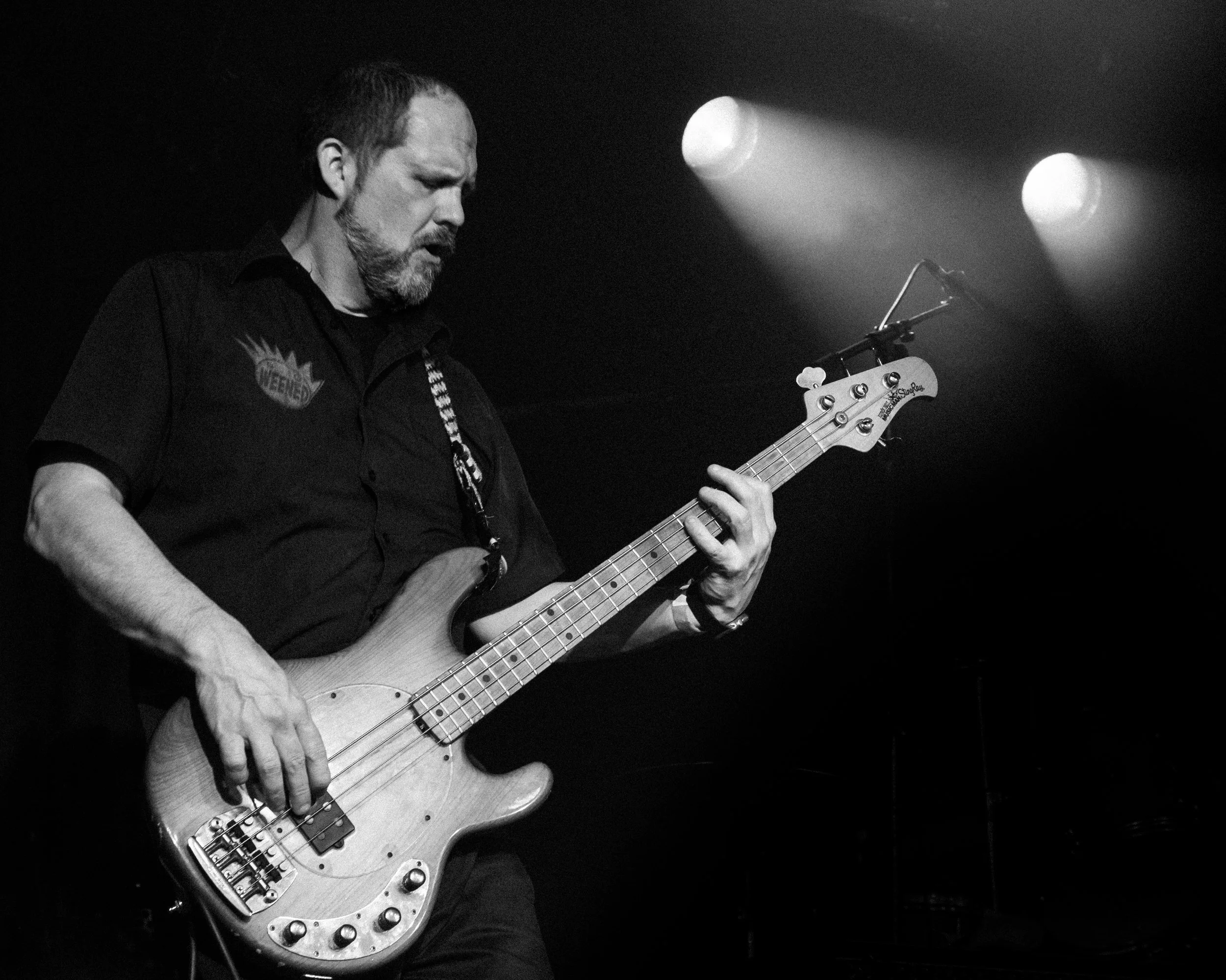 A man playing a bass guitar on stage with spotlights overhead, black and white photo