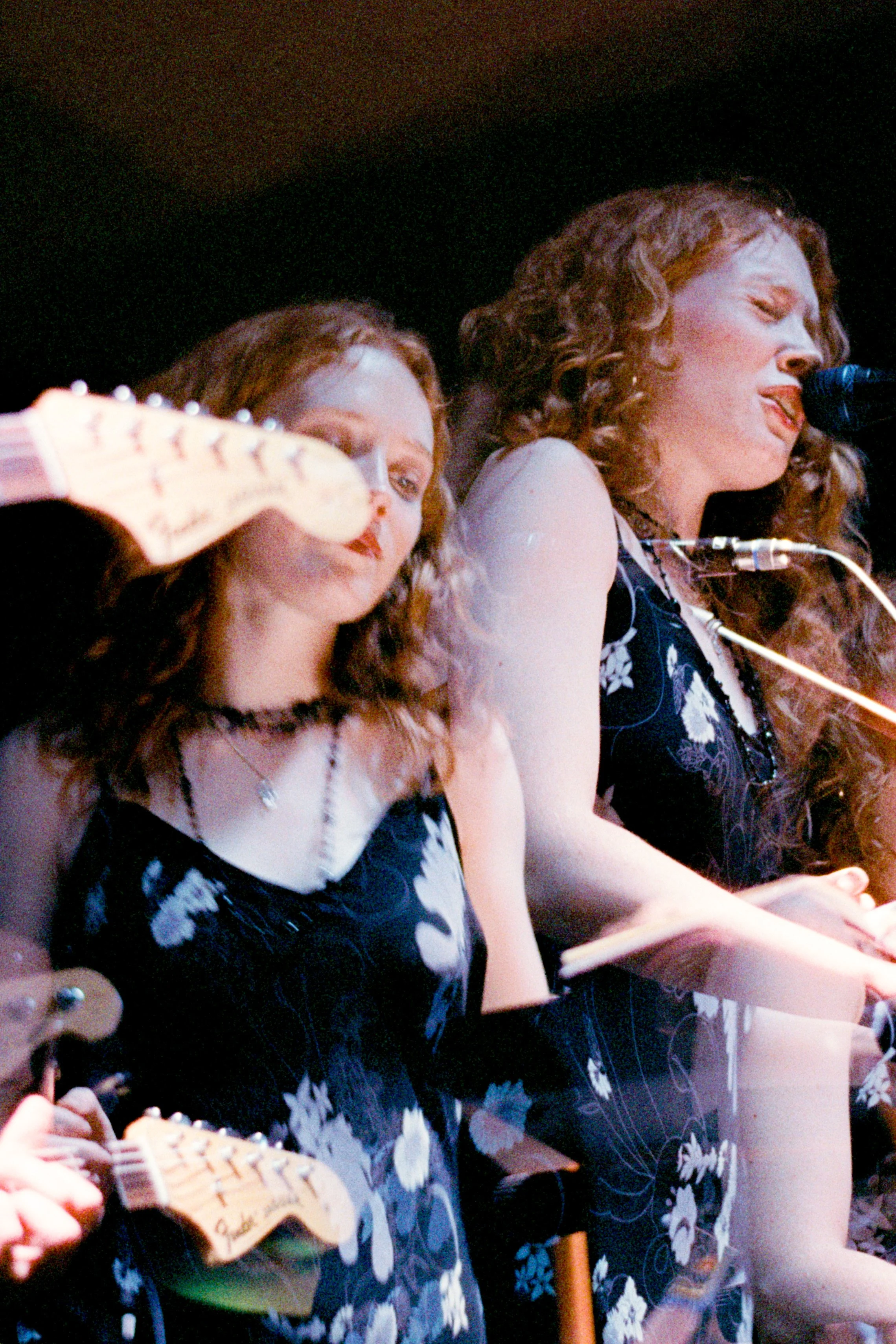 Two women performing on stage, singing into a microphone, wearing matching black dresses with white floral patterns, with a guitarist partially visible in the foreground.