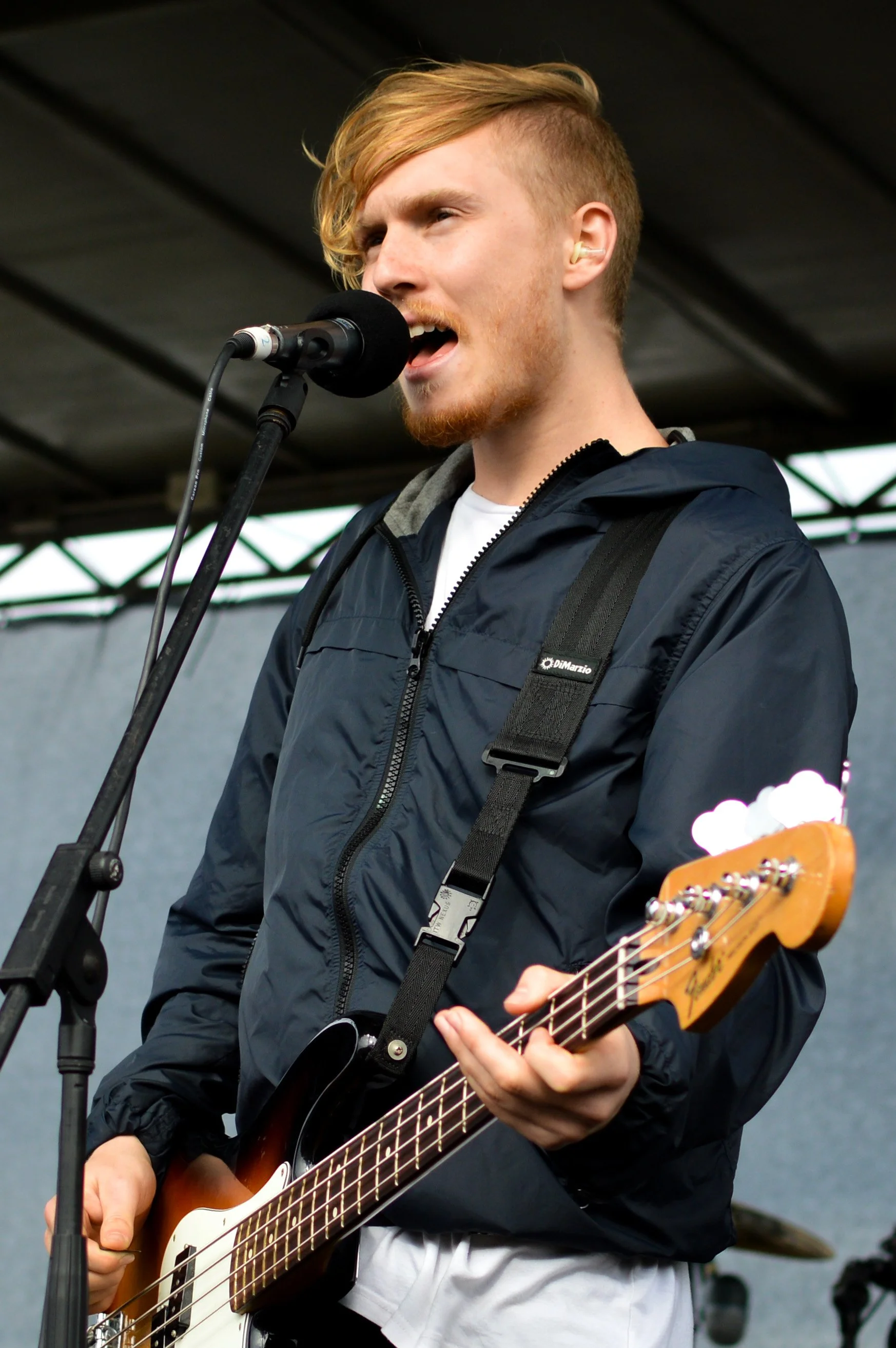 A young man performing on stage, singing into a microphone and playing an electric bass guitar.