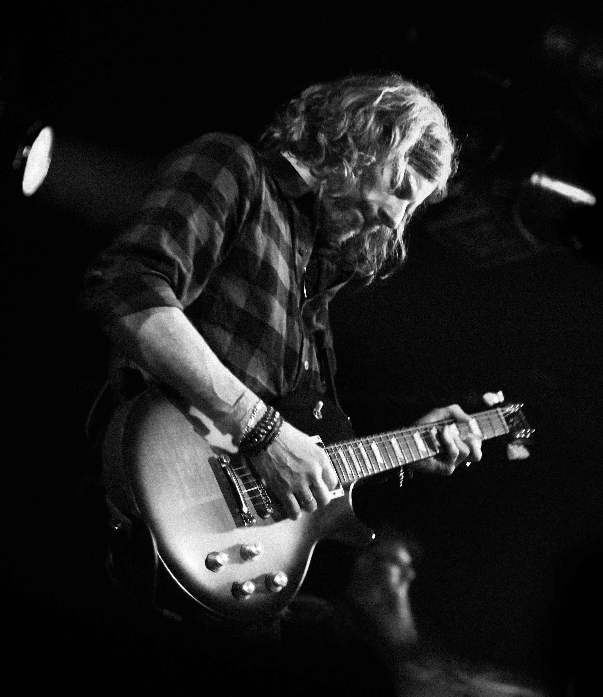 Black and white photo of a man playing an electric guitar on stage, with long hair, a beard, and wearing a checked shirt.