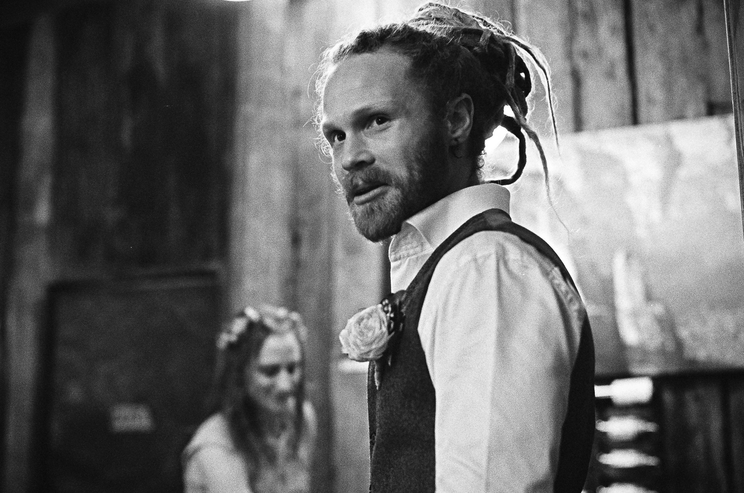 A man with dreadlocks and a beard, wearing a white shirt and vest with a flower on his lapel, looks over his shoulder in a rustic setting. A woman with braided hair and a flower crown is in the background, smiling.