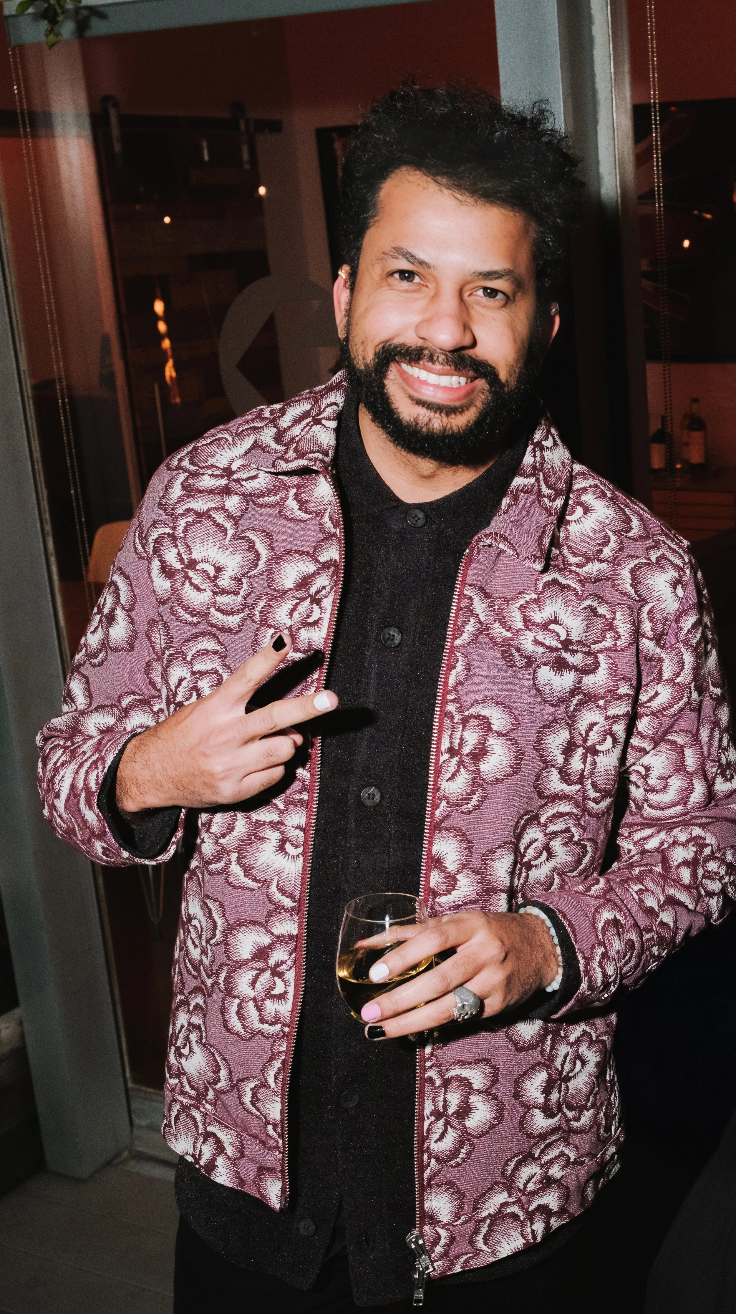 Man with curly dark hair and beard smiling, wearing a pink floral patterned jacket, holding a glass of white wine, making a peace sign with his right hand, indoors at a social event.