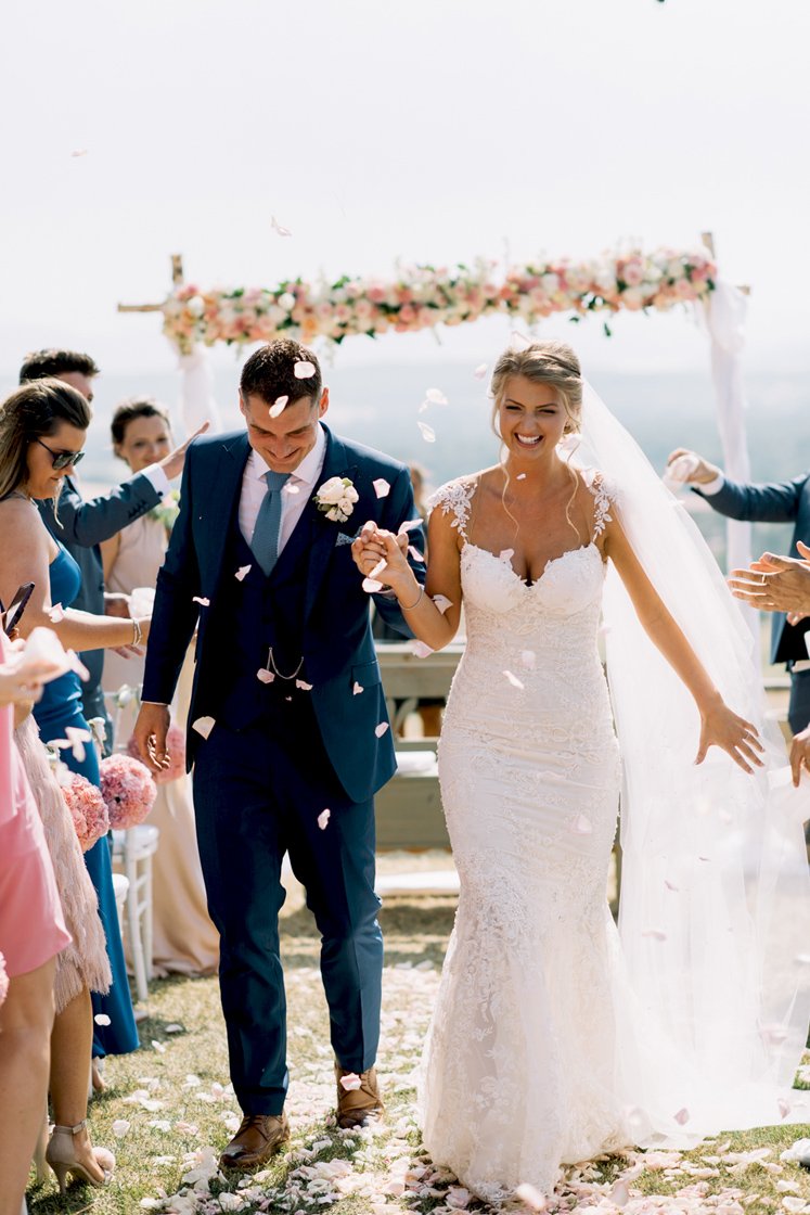 Smiling bride and groom walk down the aisle after their ceremony at Conti di San Bonifacio, guests toss flower petals during this joyful Tuscan wedding beautifully planned by FunkyBird Weddings.