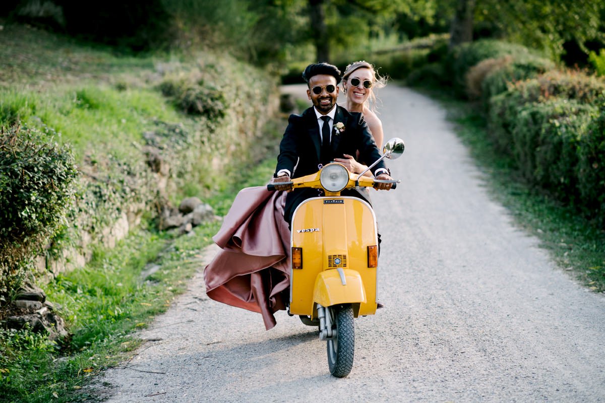 Bride and groom riding together on a yellow Vespa through the rolling hills of Val d’Orcia in Tuscany, a romantic elopement adventure in Italy, full of freedom and joy, planned by FunkyBird Weddings.