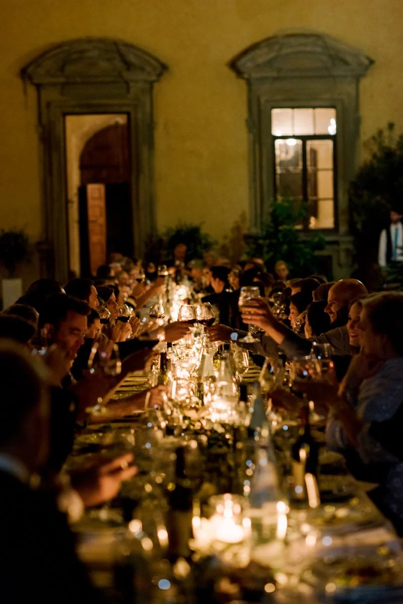 Long dinner table filled with candles during a wedding reception in Italy, guests toasting with wine at Castello di Montegufoni, a beautifully planned Tuscan wedding by FunkyBird Weddings.