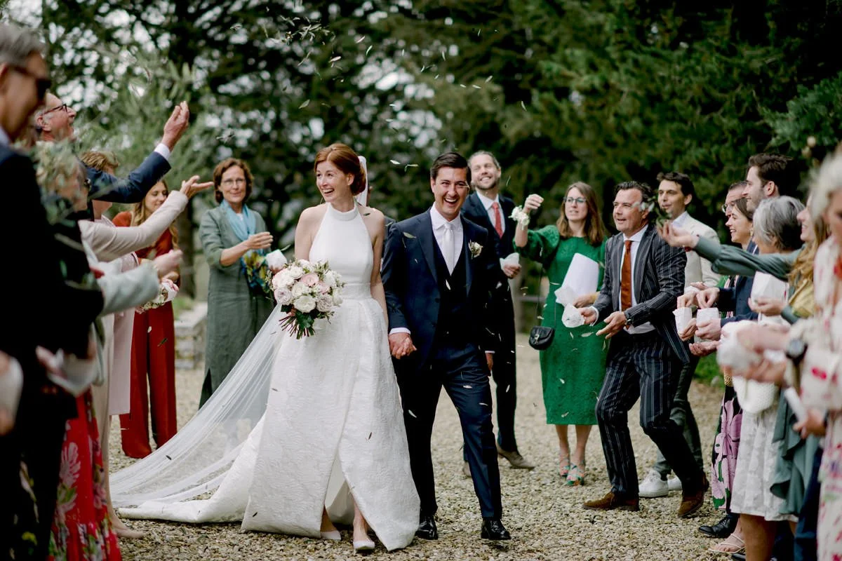 Smiling bride and groom walk away after their ceremony at Il Verreno Resort as guests toss flower petals, a relaxed, perfectly timed wedding in Tuscany coordinated by FunkyBird Weddings.
