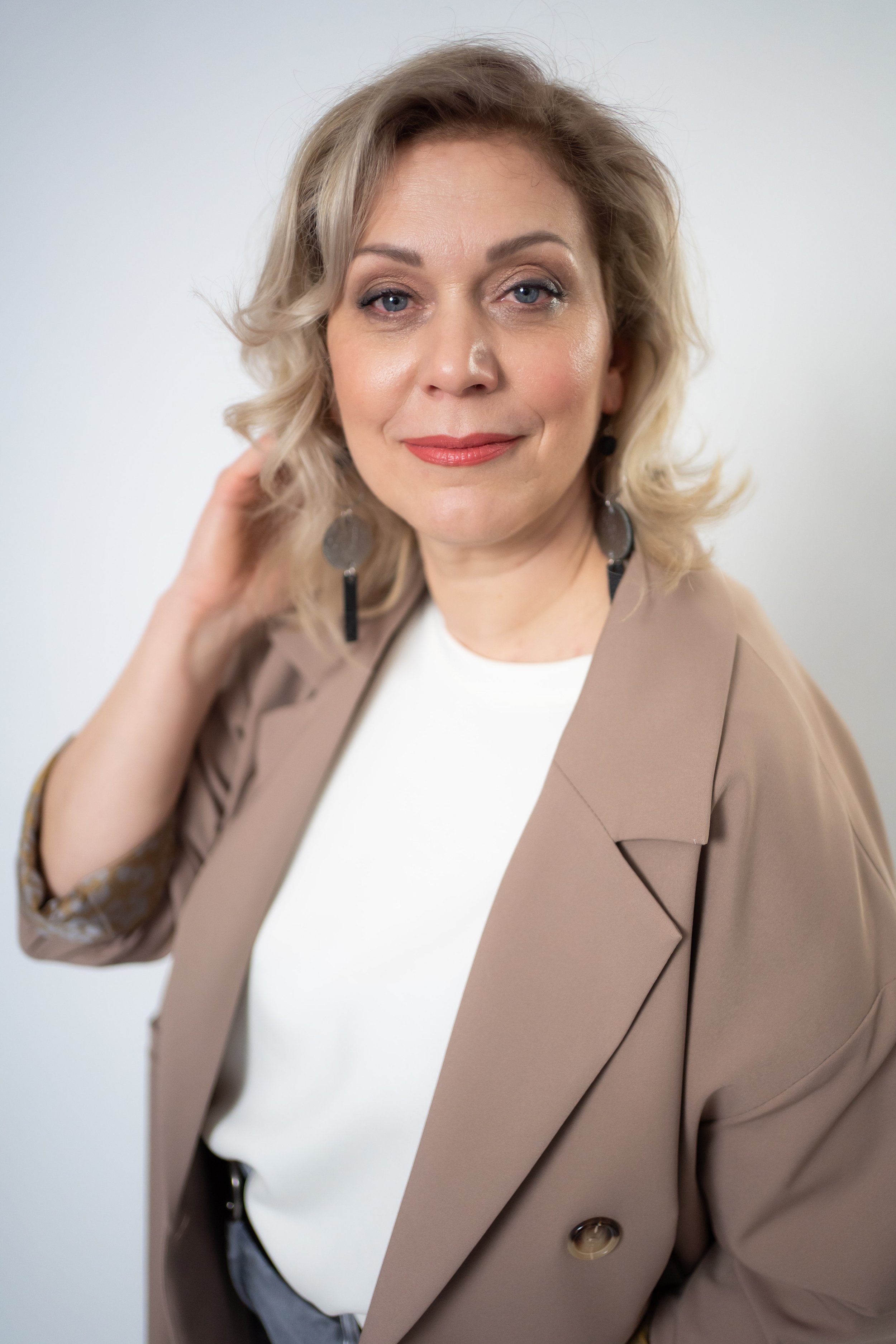 A woman with blonde, wavy hair, wearing makeup, a white top, a beige blazer, and statement earrings, posing against a light background.