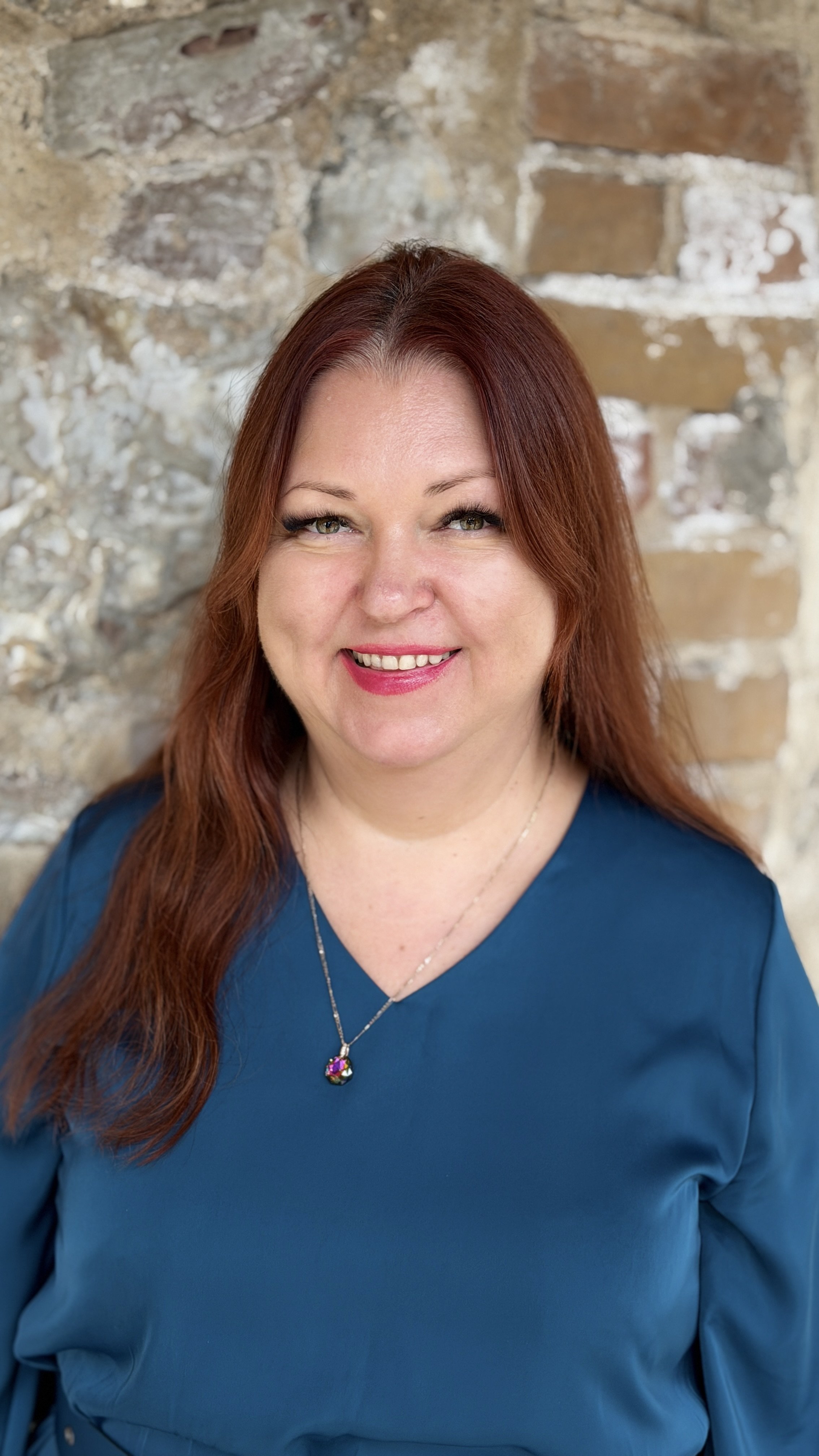 A woman with long red hair smiling, wearing a navy blue top and a pendant necklace, standing in front of a stone wall.
