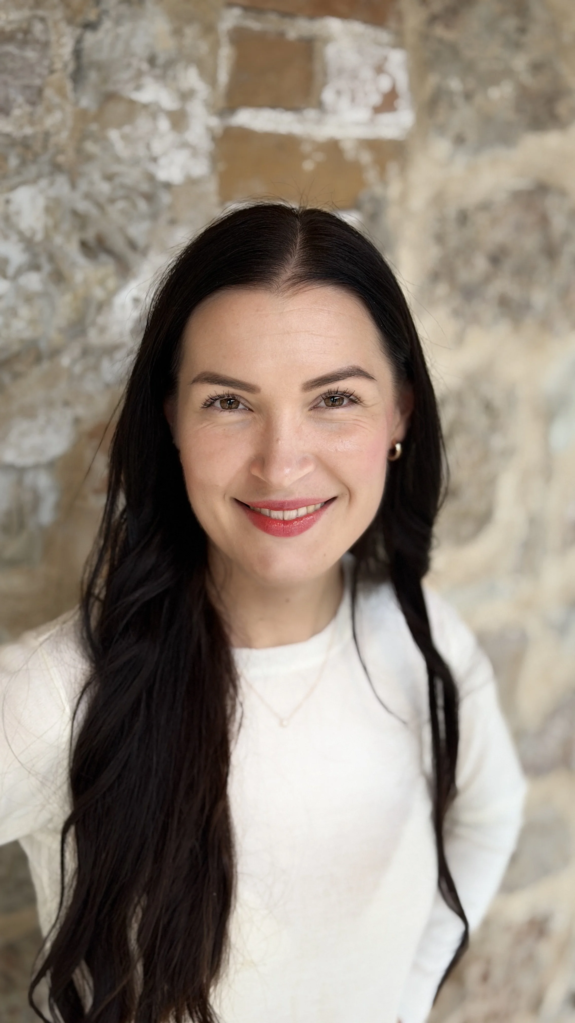 Close-up portrait of a smiling woman with long dark hair, wearing a white top, standing against a rustic brick wall.