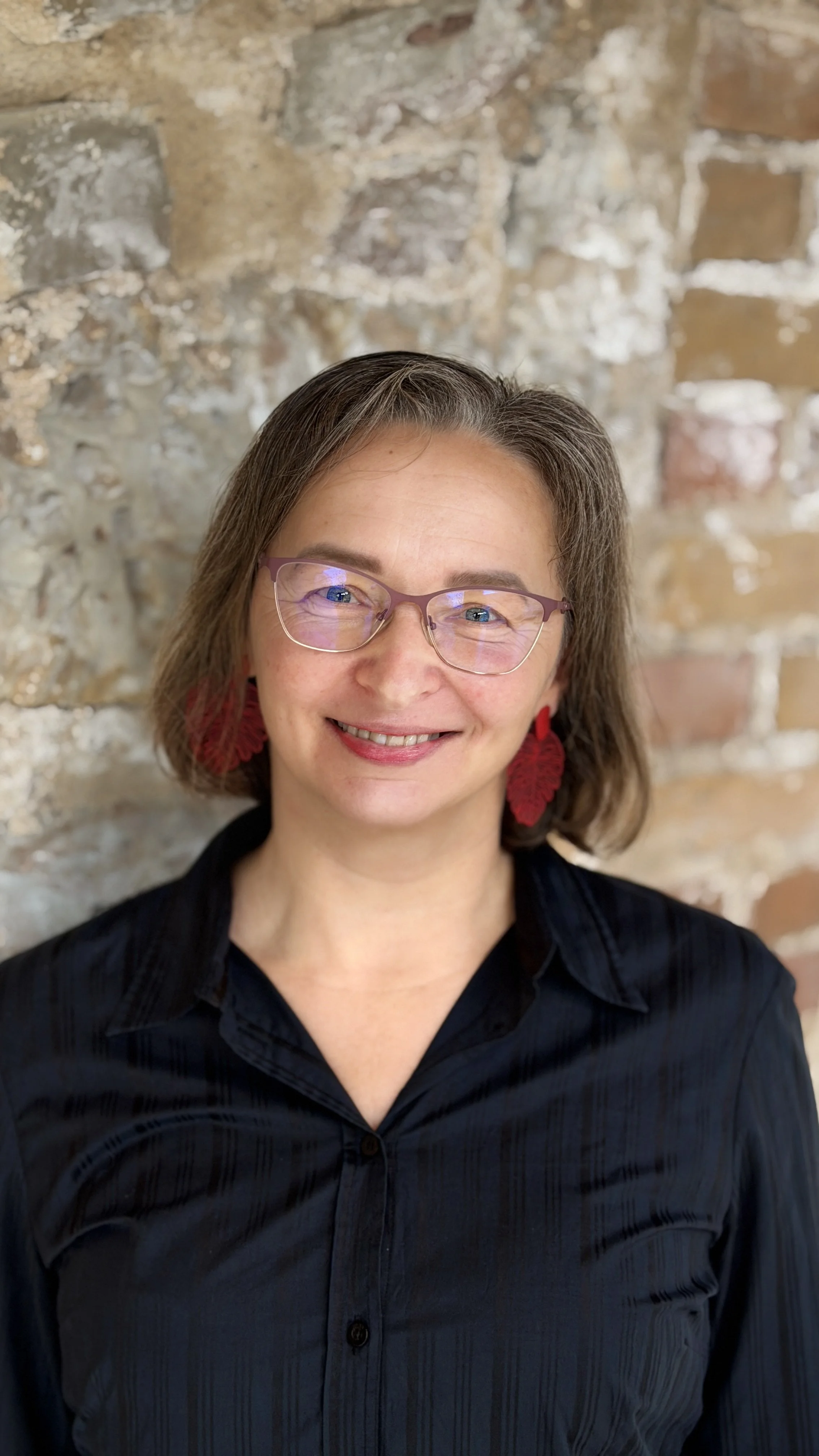 A woman with short brown hair, wearing glasses, red earrings, and a black shirt, smiling against a brick wall background.