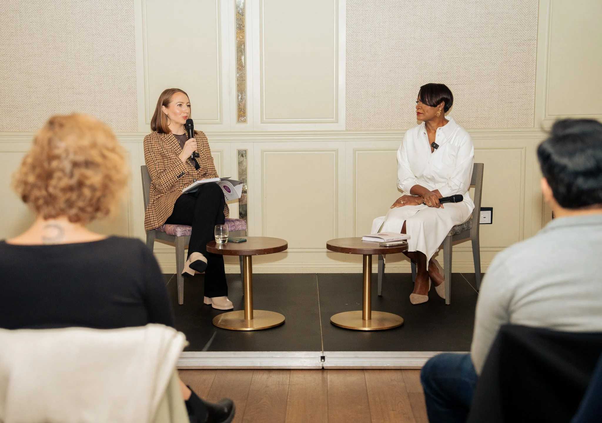 A woman speaking with a microphone during a panel discussion in a conference room with three audience members visible.