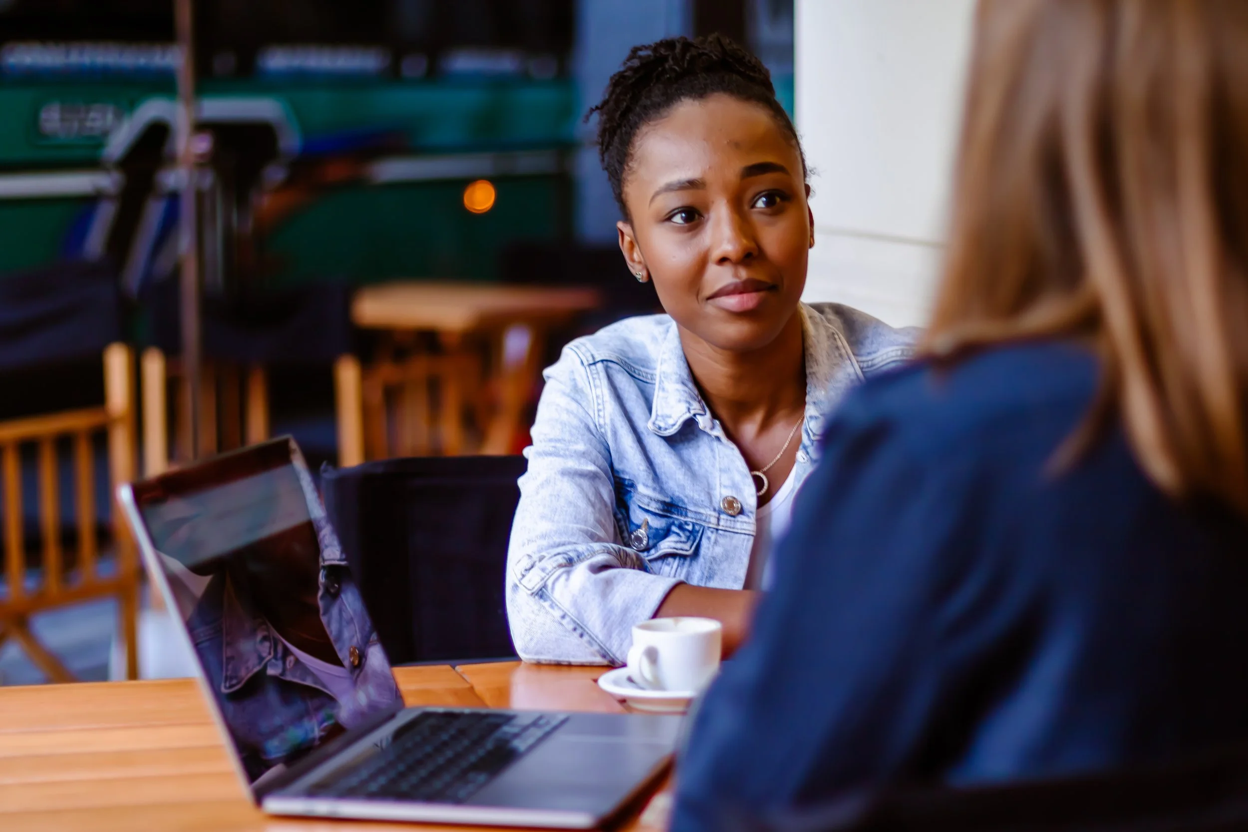 Two women sitting at a table with a laptop and a small cup; one woman with dark skin and black hair in a bun looking attentively at the other woman, who has blonde hair and is turned away from the camera, in a casual cafe setting.