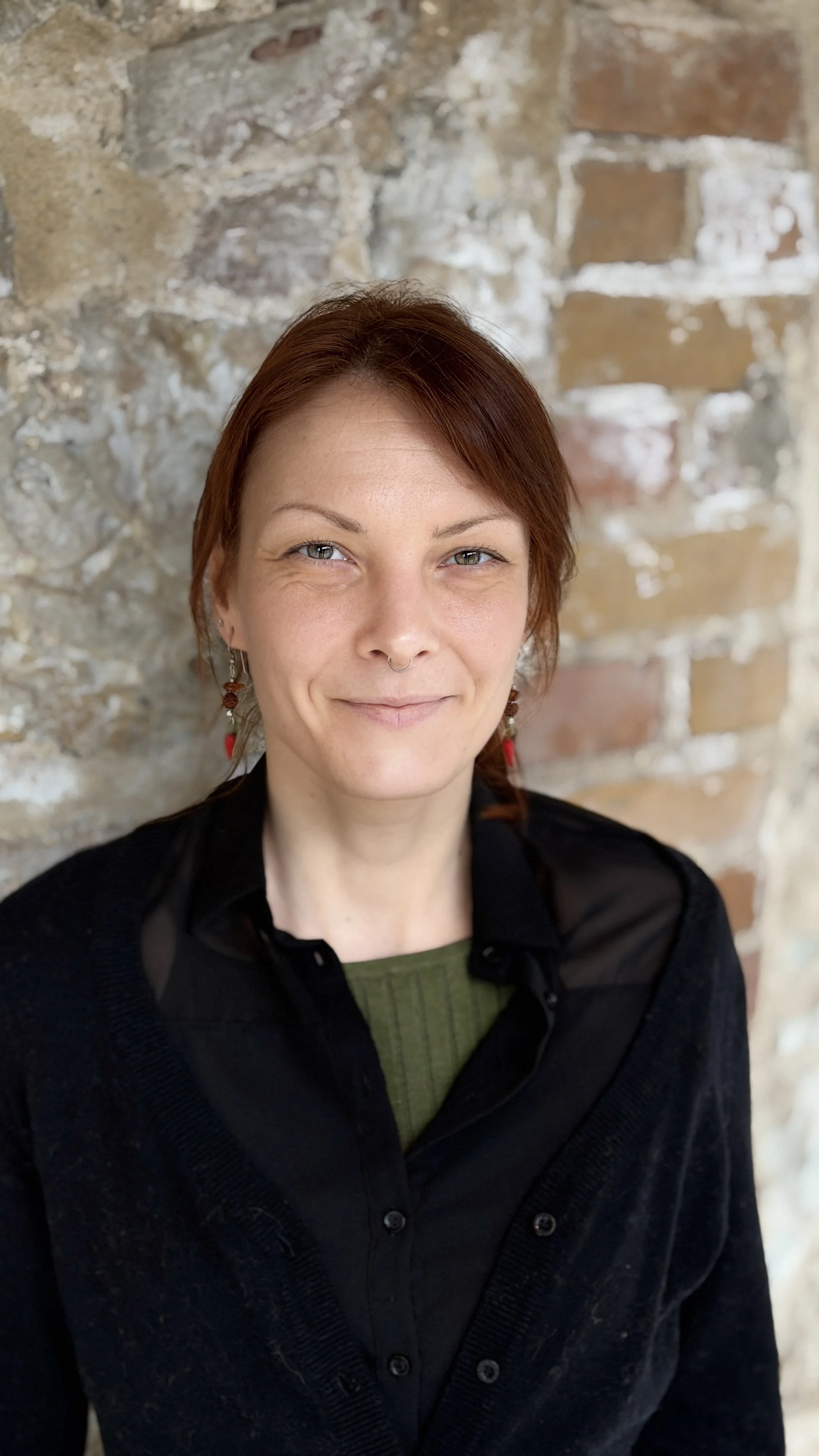 A woman with short reddish-brown hair, light skin, and light eyes, smiling slightly, standing in front of a brick wall. She is wearing earrings, a black top with sheer shoulder details, and a green shirt underneath.