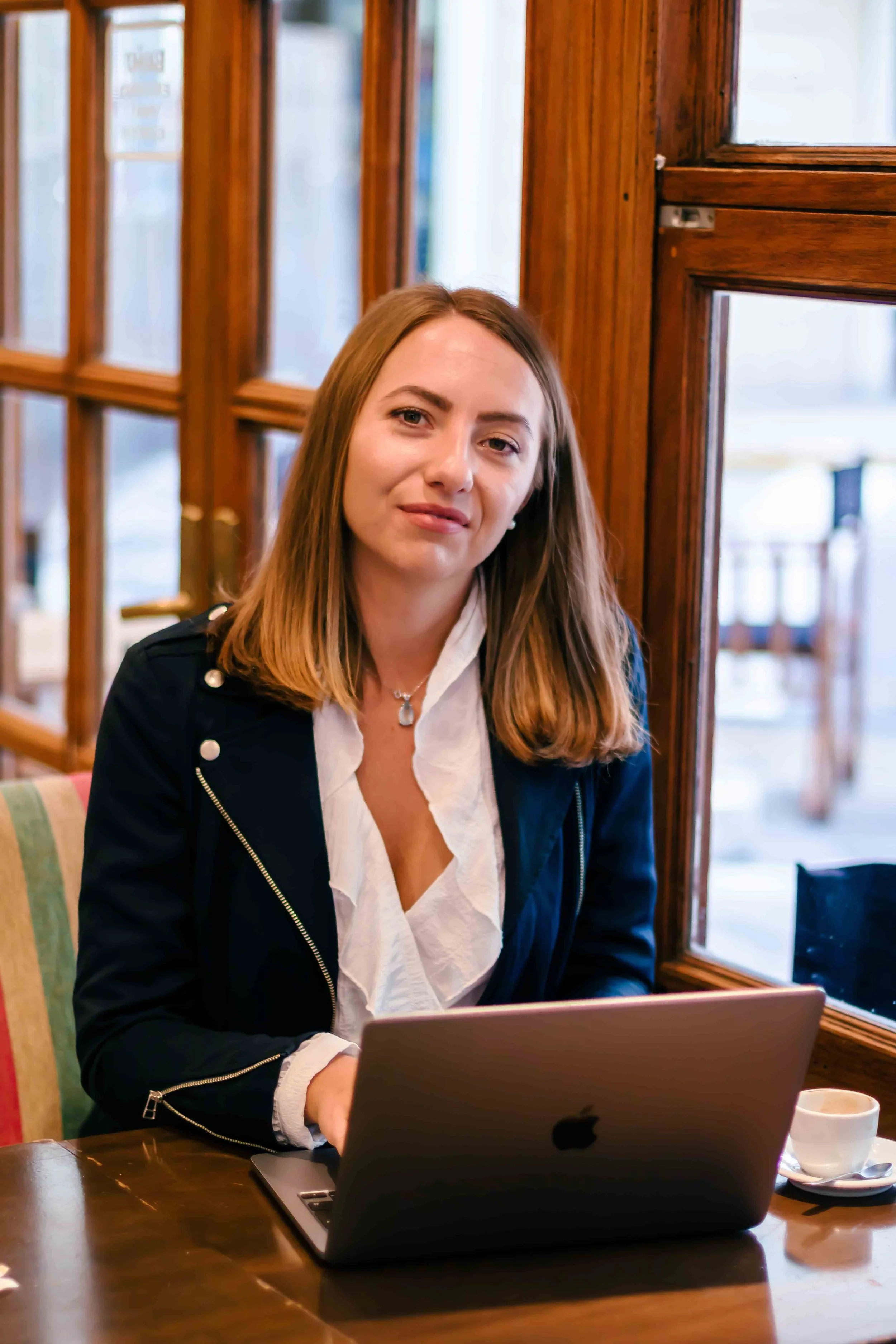 A woman sitting at a wooden table in a cozy cafe, working on a laptop with a cup of coffee nearby, by a window with wooden framing.