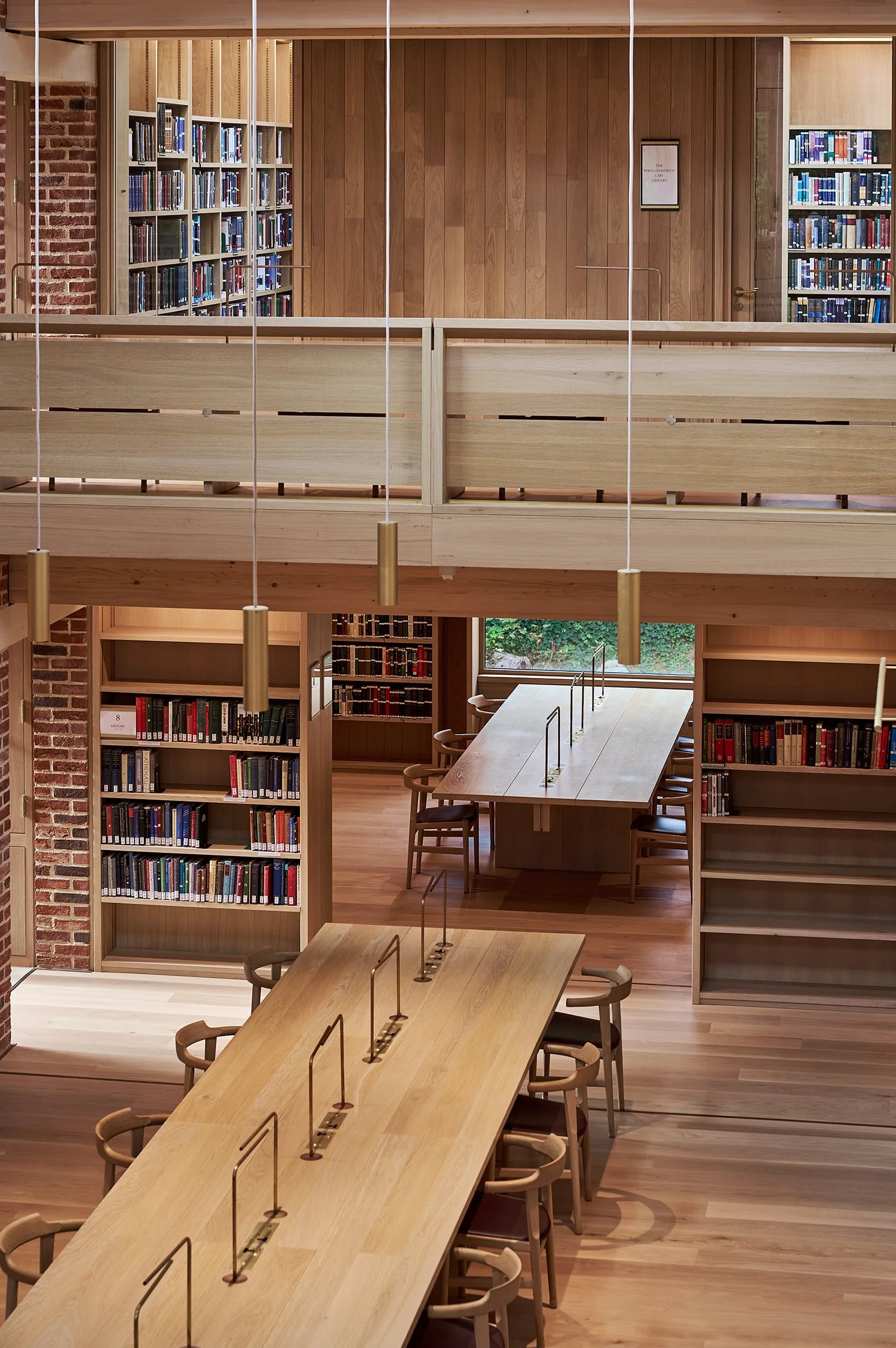 Interior of a modern library with wooden tables, chairs, and bookshelves. The design features wooden paneling and vertical lighting fixtures. Bookshelves are filled with books, and a large window offers natural light.